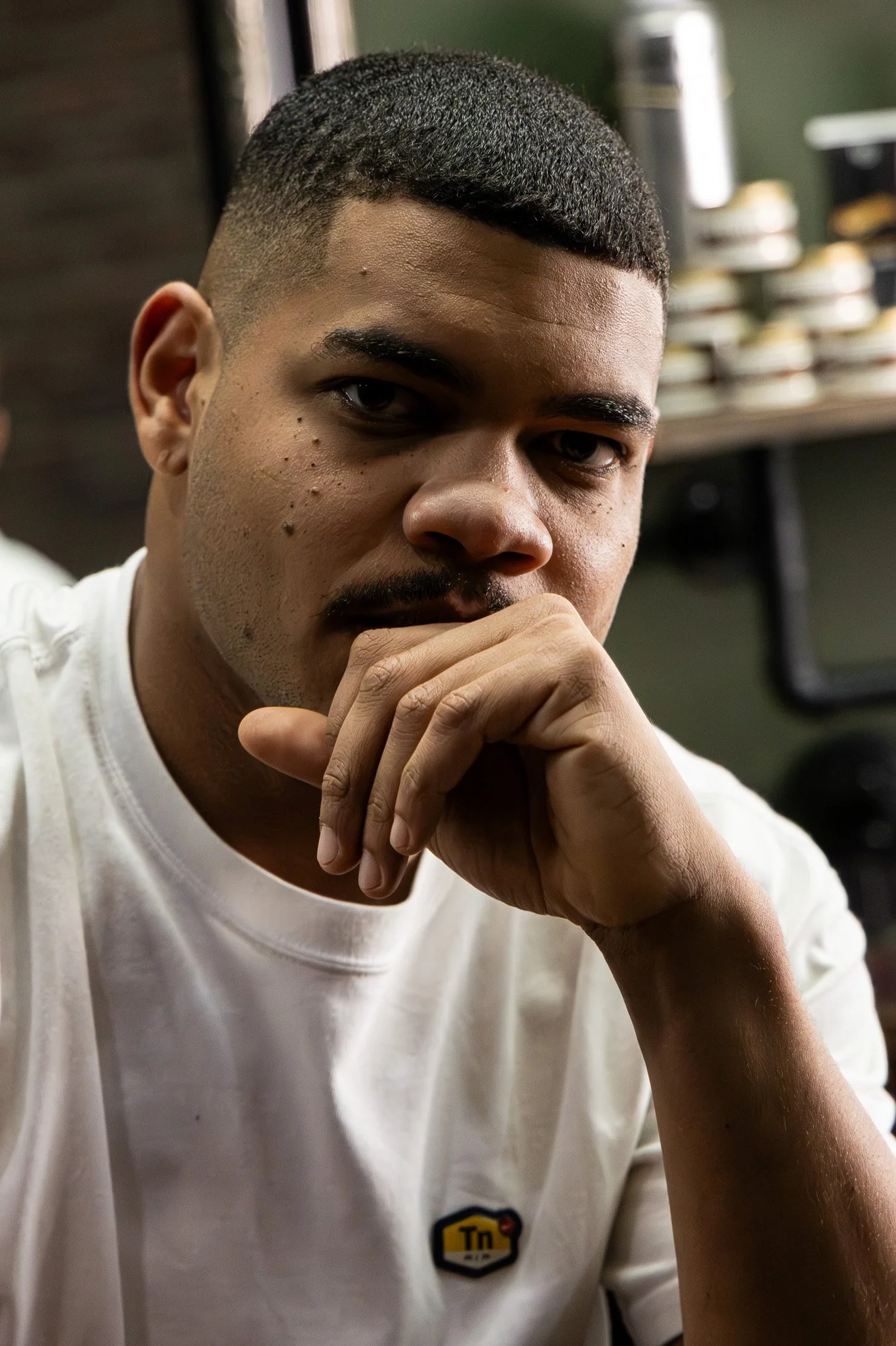 Close-up of a young man with short dark hair and facial hair, wearing a white T-shirt, resting his chin on his hand with a thoughtful expression. Shelves with various items are in the background.