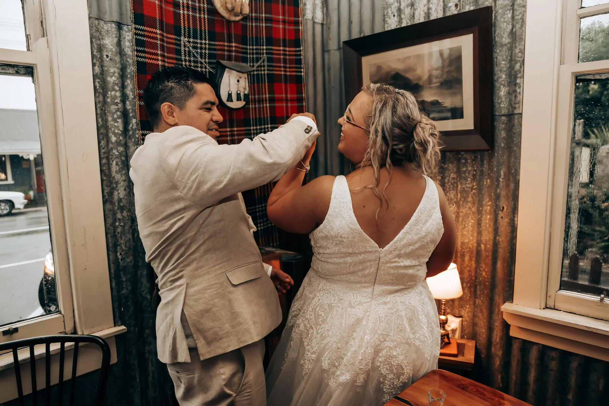 A man and woman, possibly a bride and groom, holding hands and smiling at each other indoors near windows, with rustic decor including a tartan cloth and forest artwork on the wall.