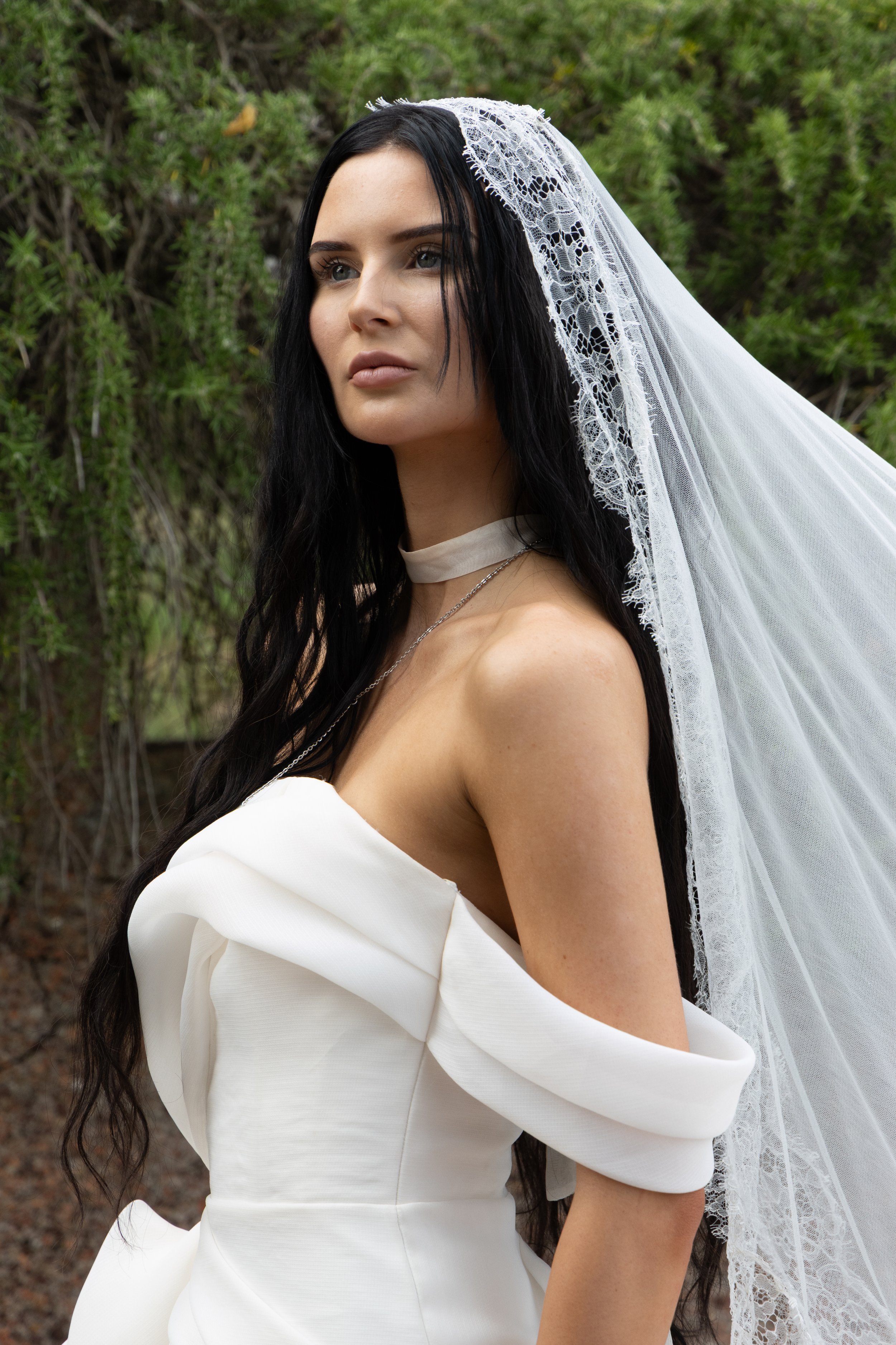 A woman with long black hair wearing a white off-shoulder dress and a lace veil outdoors.