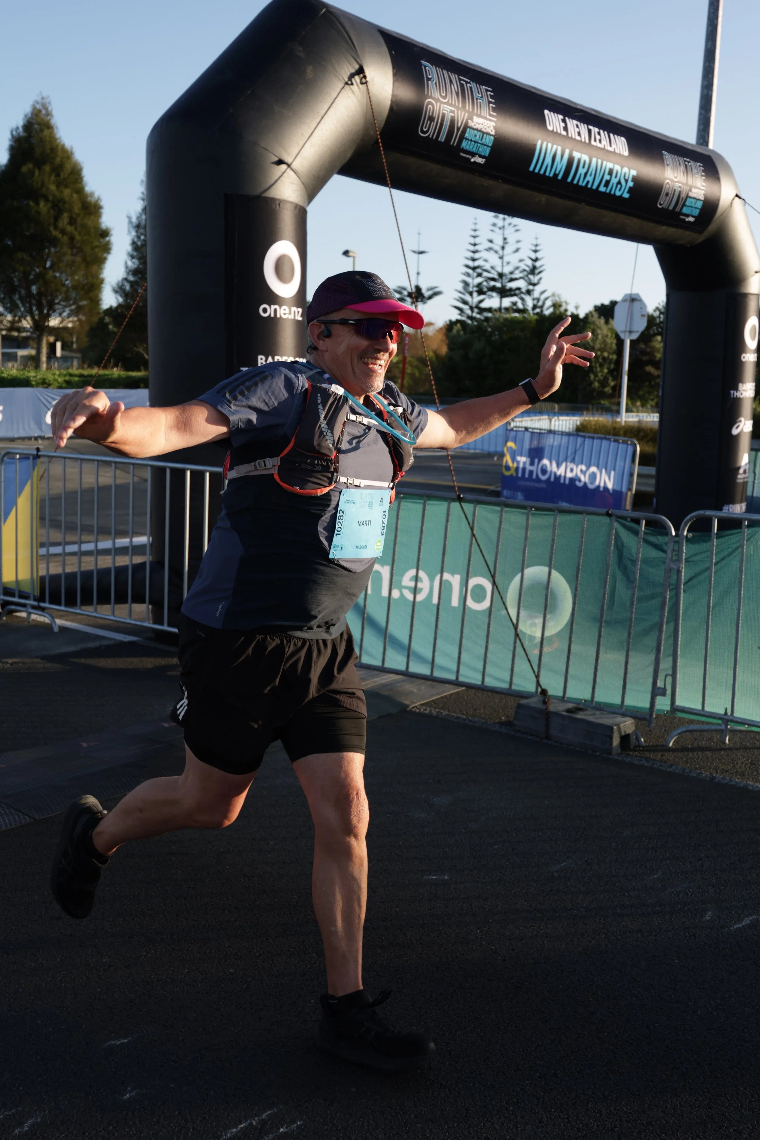 Male marathon runner crossing the finish line, smiling with arms outstretched, wearing black shorts, gray t-shirt, sunglasses, hat, and a hydration pack, at an outdoor race event with a large inflatable arch and banners.