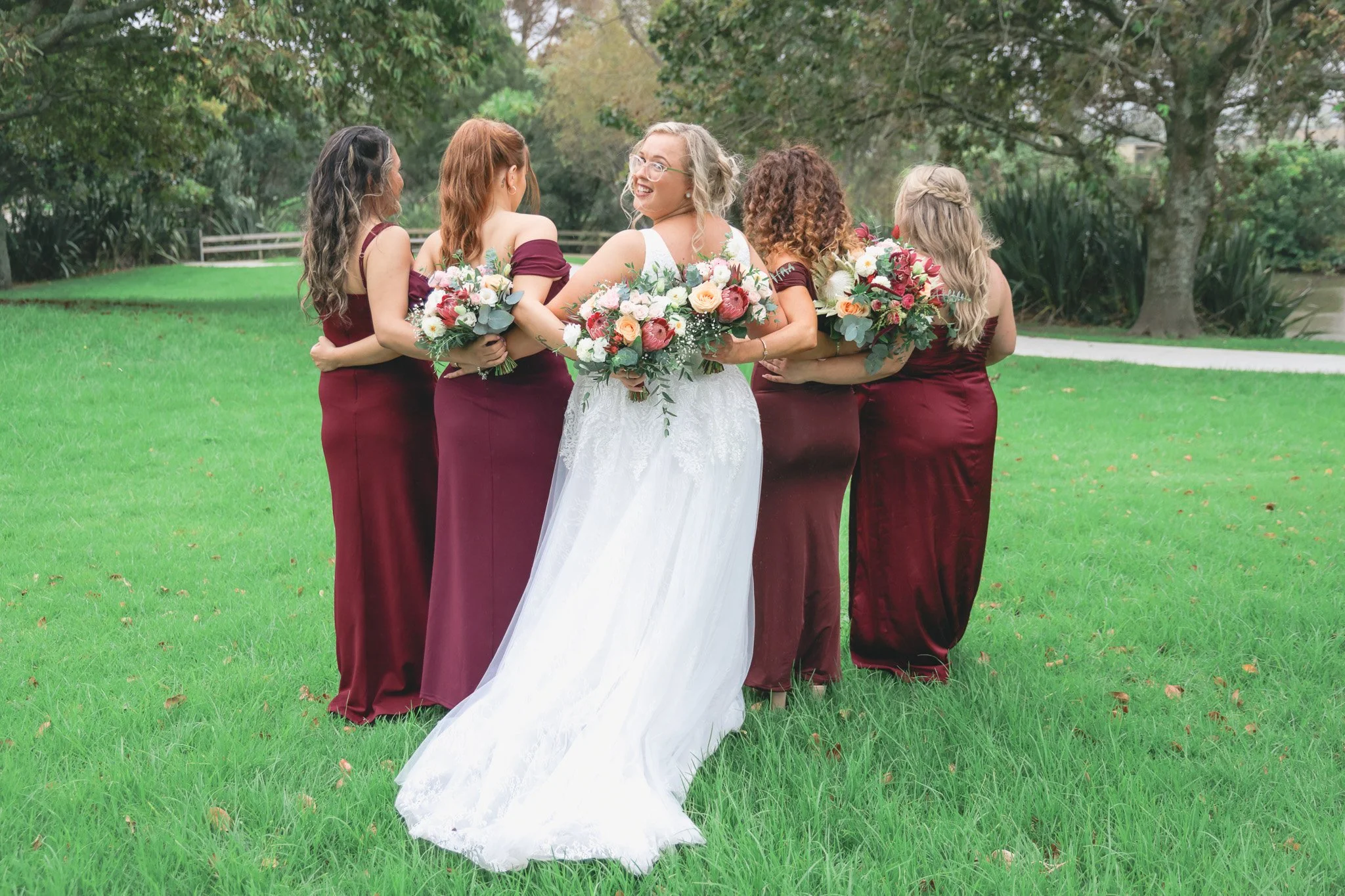 A bride in a white wedding dress holding a bouquet of flowers, surrounded by five women in burgundy dresses holding similar bouquets, standing on a grassy area with trees in the background, engaging in conversation at a wedding.