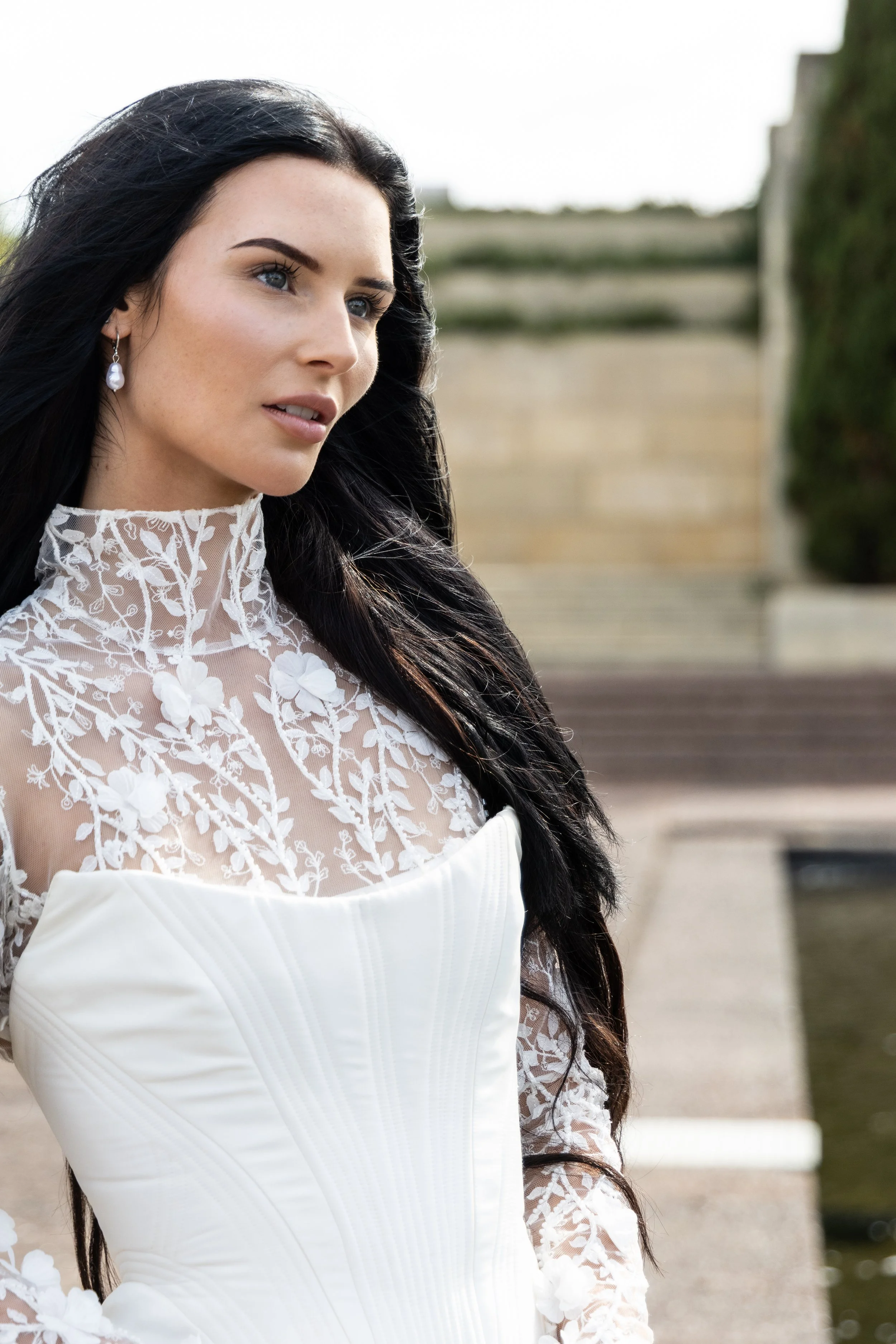 A woman with long dark hair wearing a white dress with lace floral details and a pearl earring, standing outdoors near stairs and greenery.