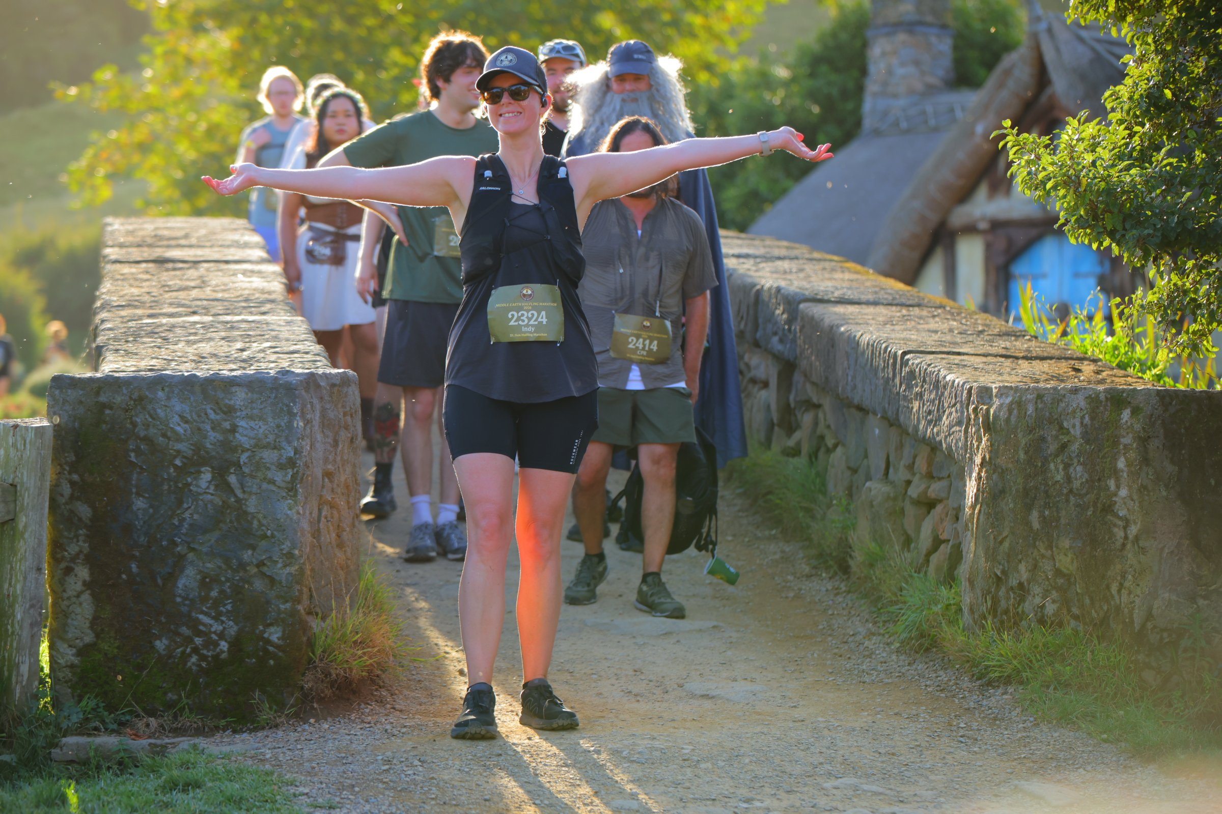 A group of people participating in a marathon or race walking on a stone bridge during sunset. The woman in front has her arms outstretched, smiling, wearing sunglasses, a black sleeveless top, shorts, and a bib with the number 2324. The group includ