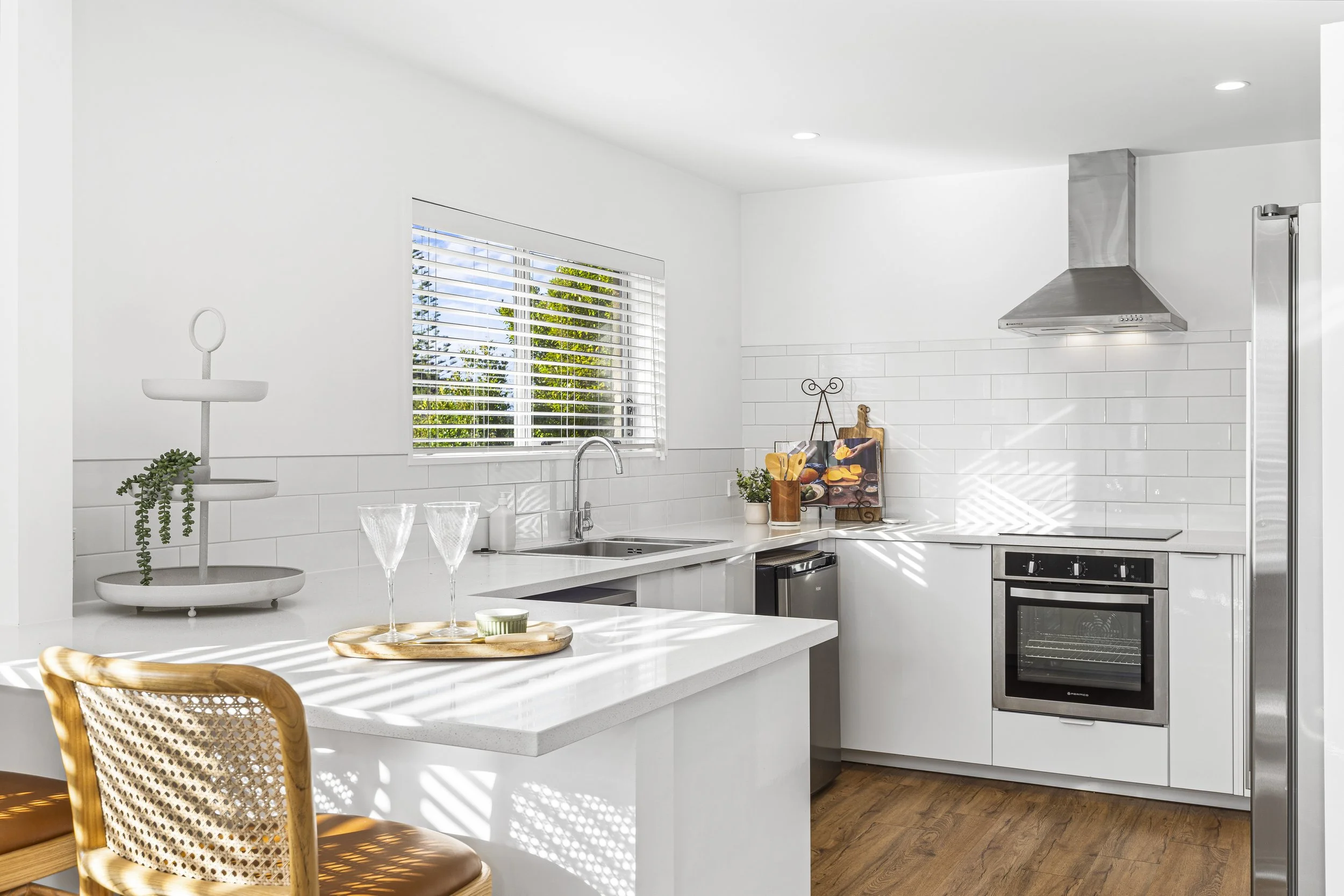 Modern kitchen with white cabinets, stainless steel appliances, a window with blinds, a white countertop, and wooden chairs. Decor includes plants and a tray with glasses and food.