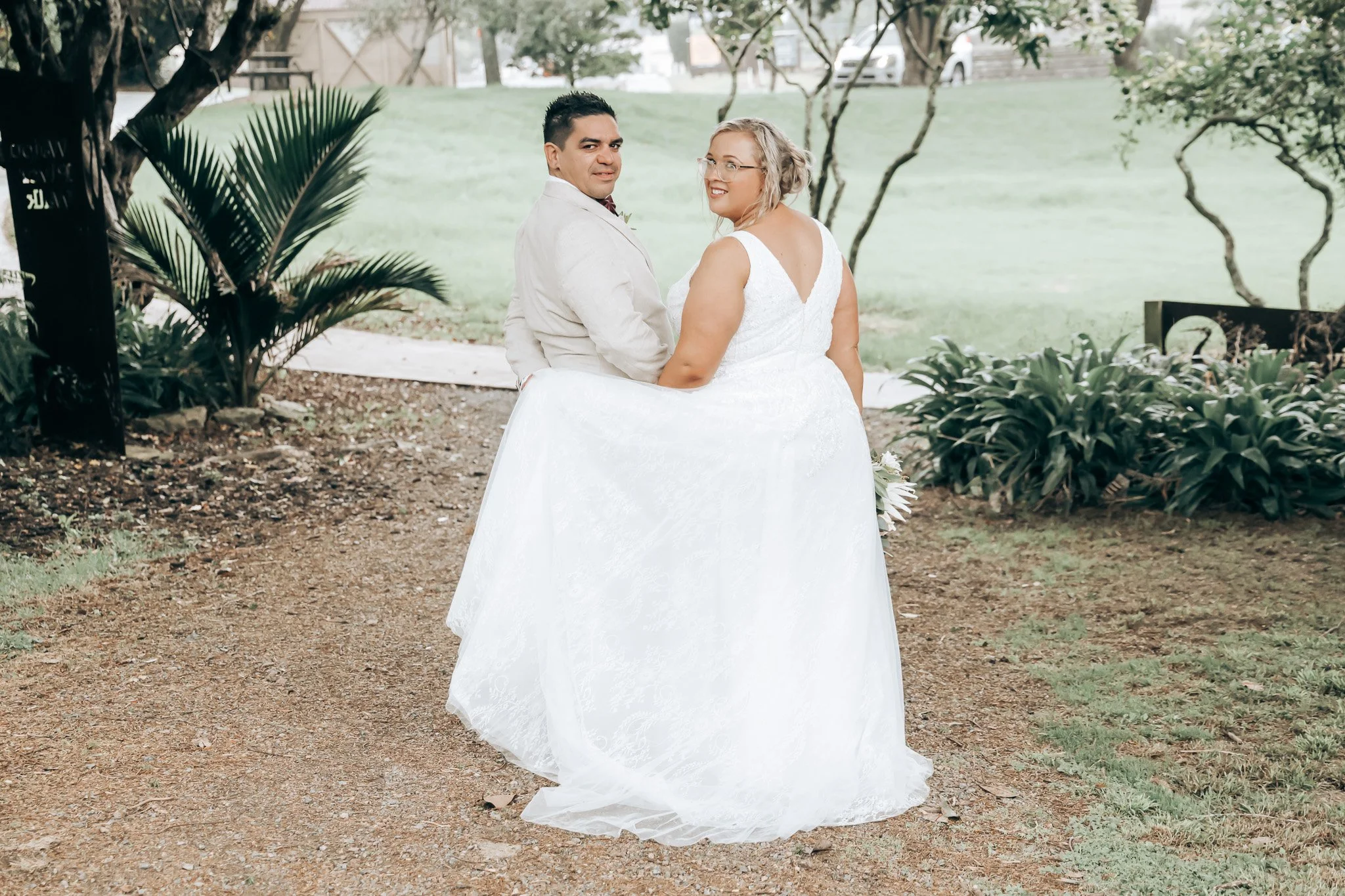 A bride and groom sitting outdoors on a white bench during their wedding, surrounded by greenery and trees.
