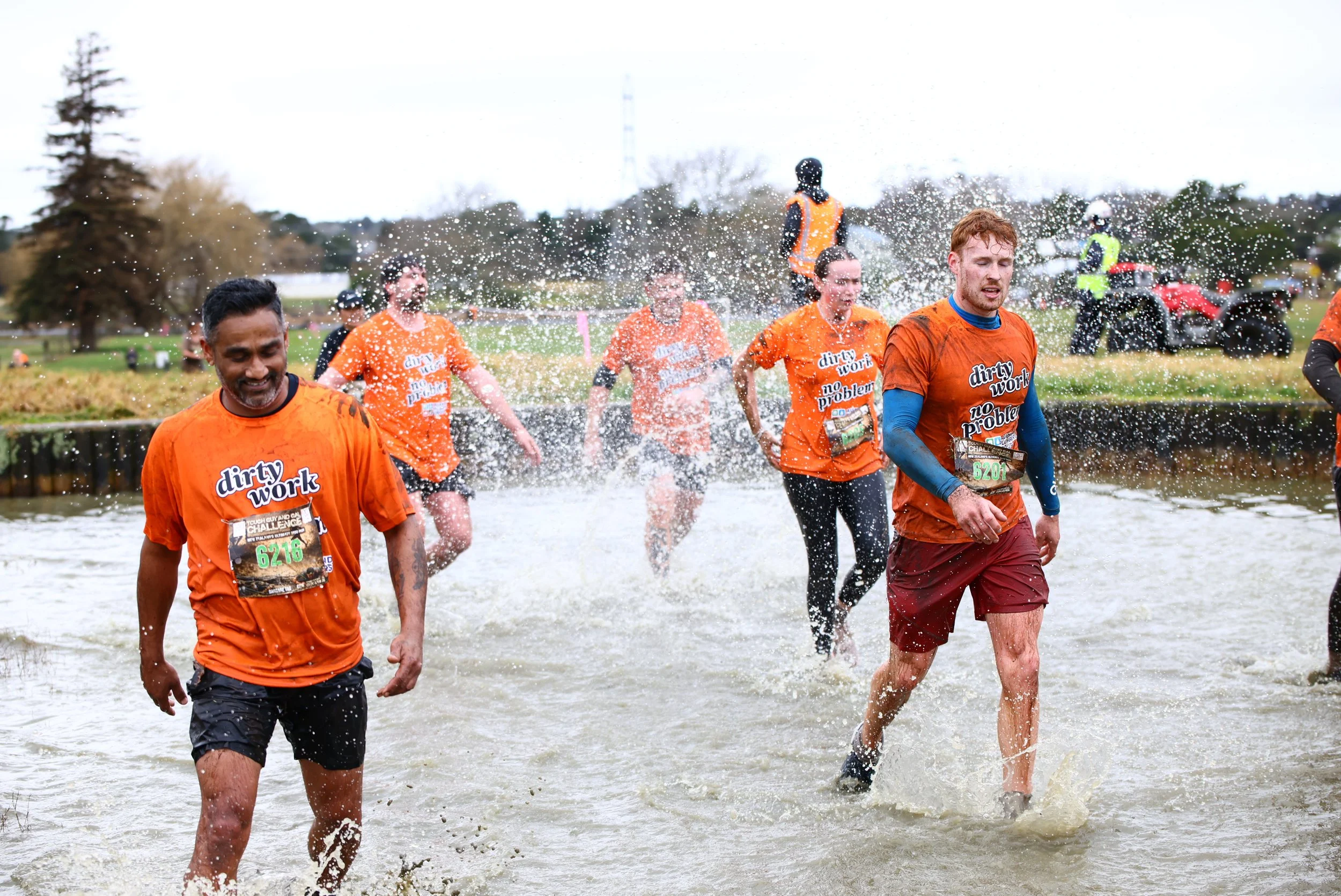 Group of runners in orange shirts participating in an outdoor event, crossing a water obstacle during a muddy race.