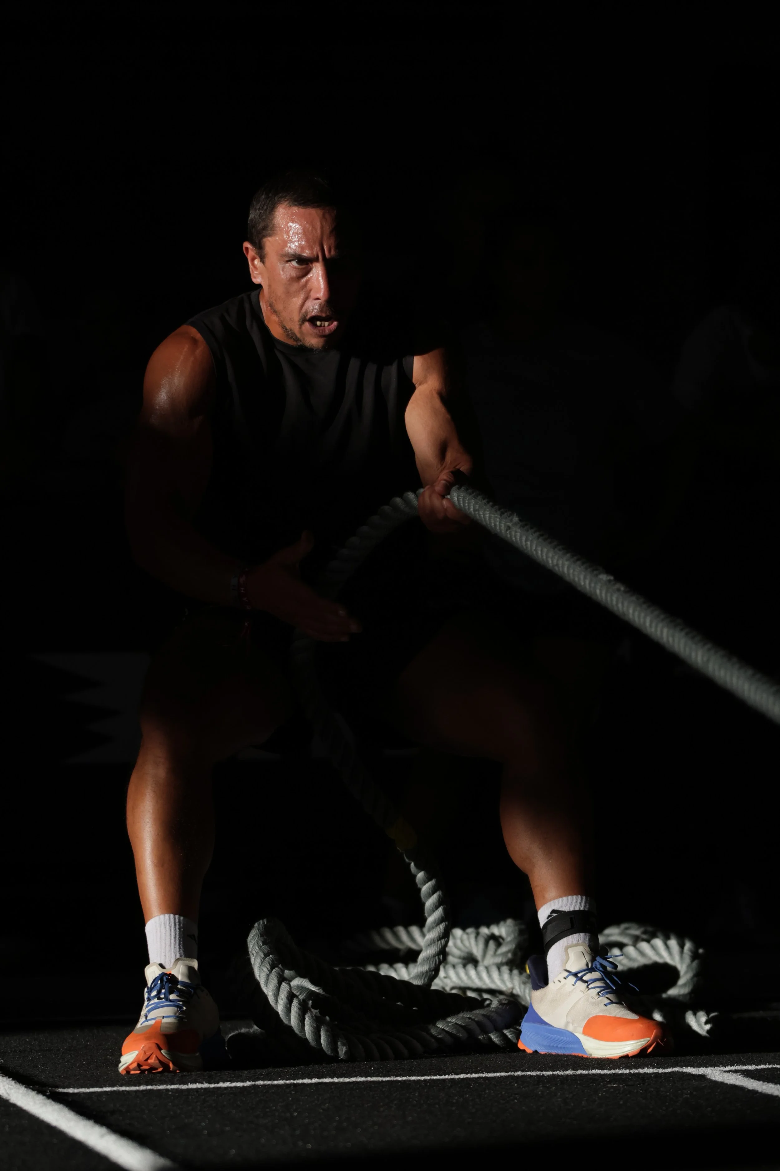 Man in athletic clothing appears to be in the middle of a workout or sports training, pulling on battle ropes in a dark gym setting.