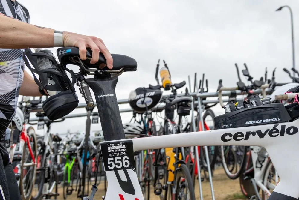 Person adjusting the seat on a Cervélo racing bicycle at a triathlon event, with multiple bikes of various brands parked on racks in the background