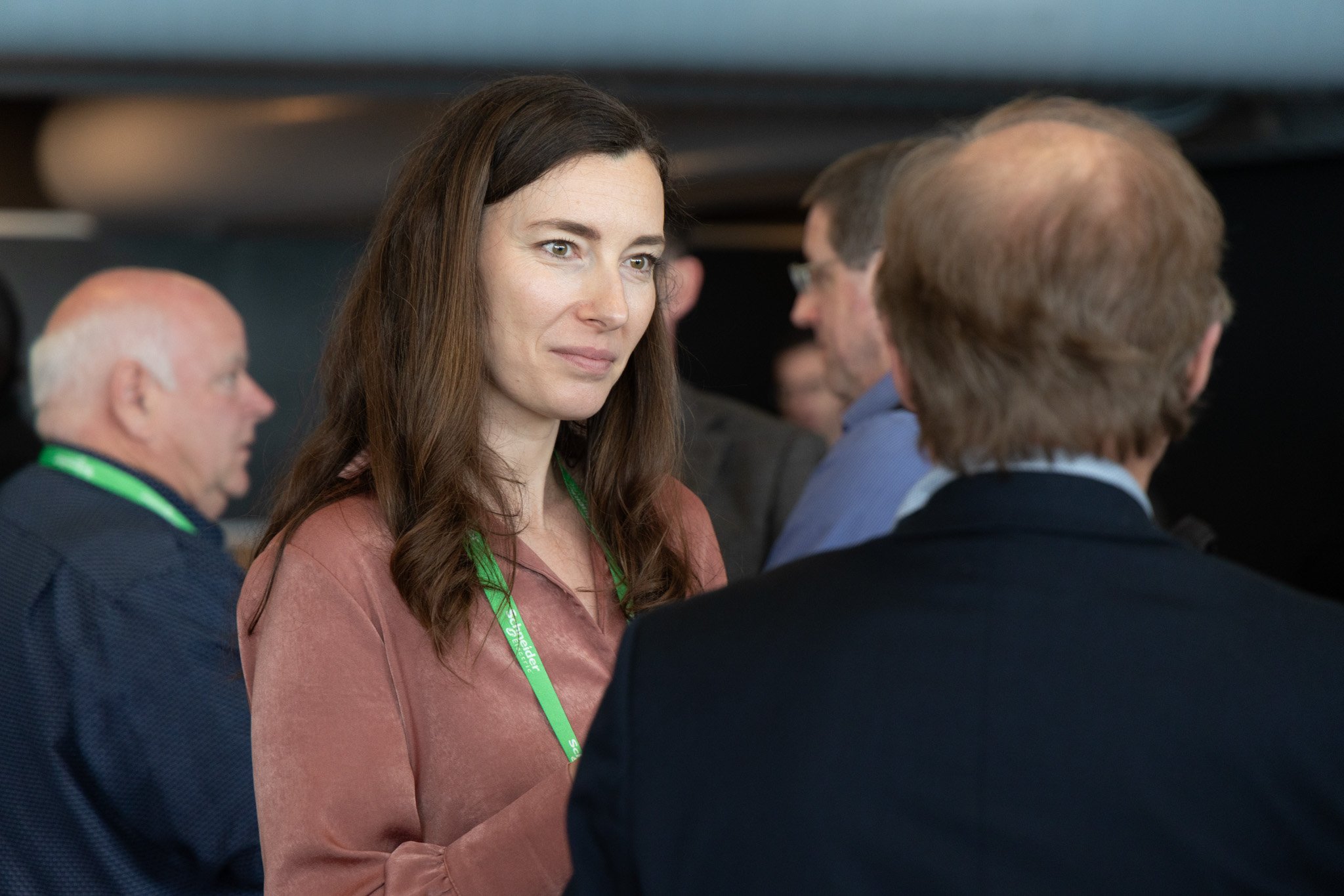 A young woman with long brown hair and light eyes, wearing a reddish-brown top and a green lanyard, is engaged in conversation with an older man with glasses and graying hair. In the background, other people are also conversing, suggesting a professi