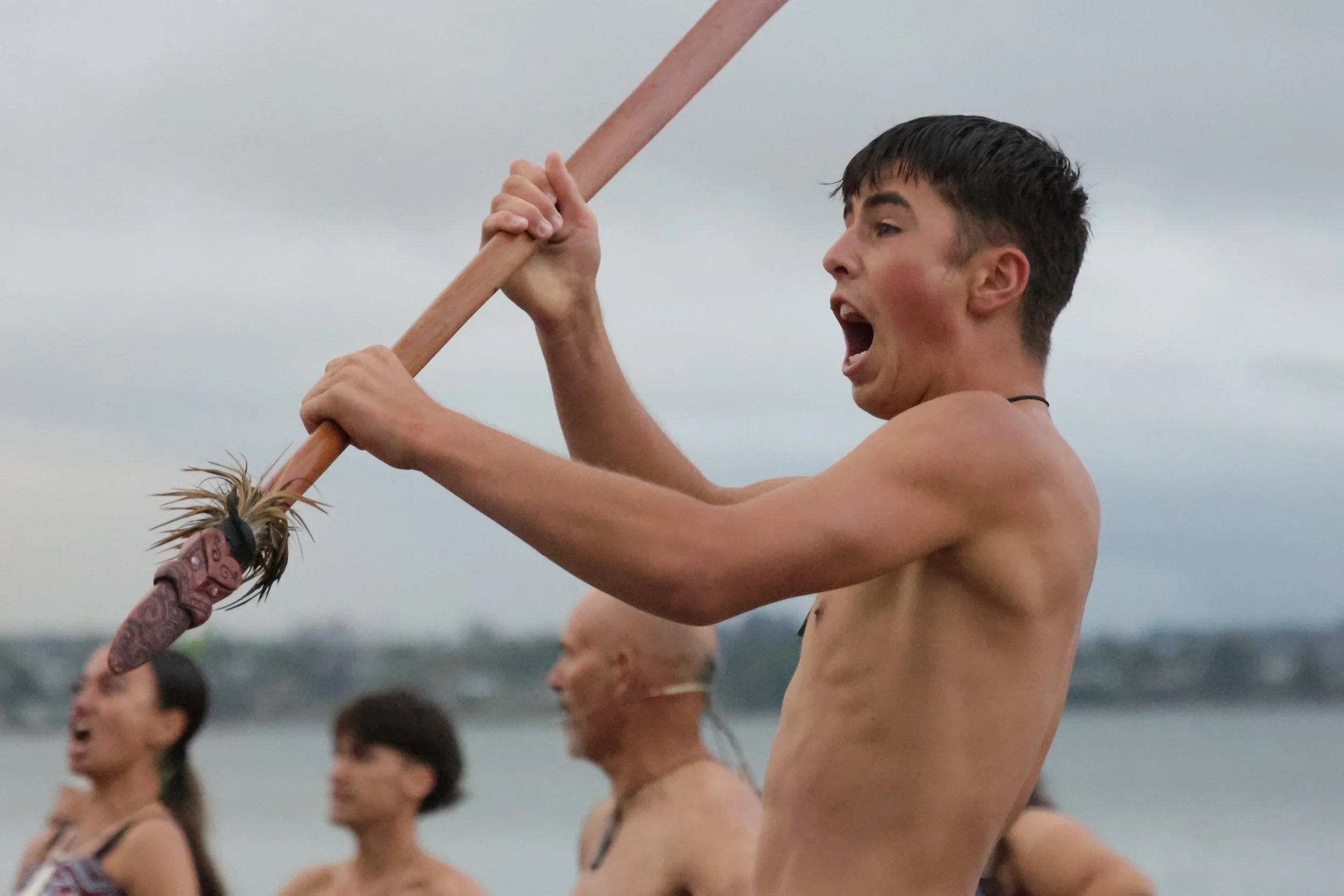 Young individuals participating in a traditional cultural dance called Haka, performing with wooden sticks on a beach during daytime.
