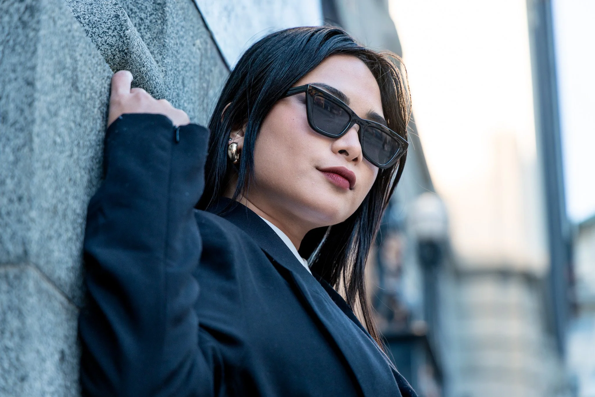 A woman with dark hair, wearing black sunglasses and earrings, leaning against a concrete wall outdoors.