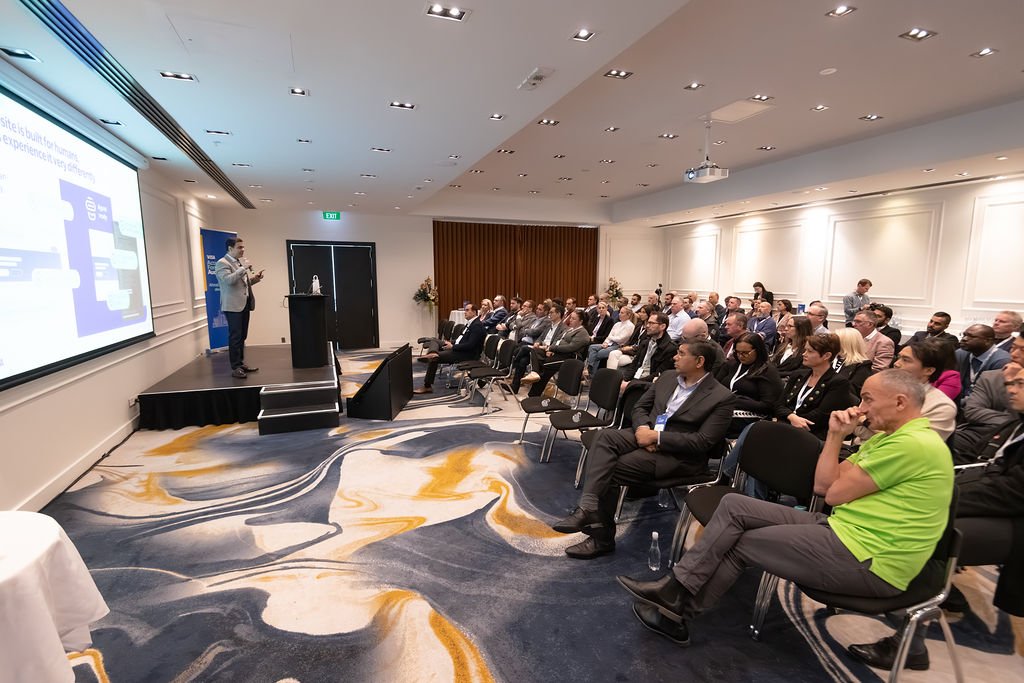 Business conference with speaker on stage presenting to an audience in a hotel ballroom.