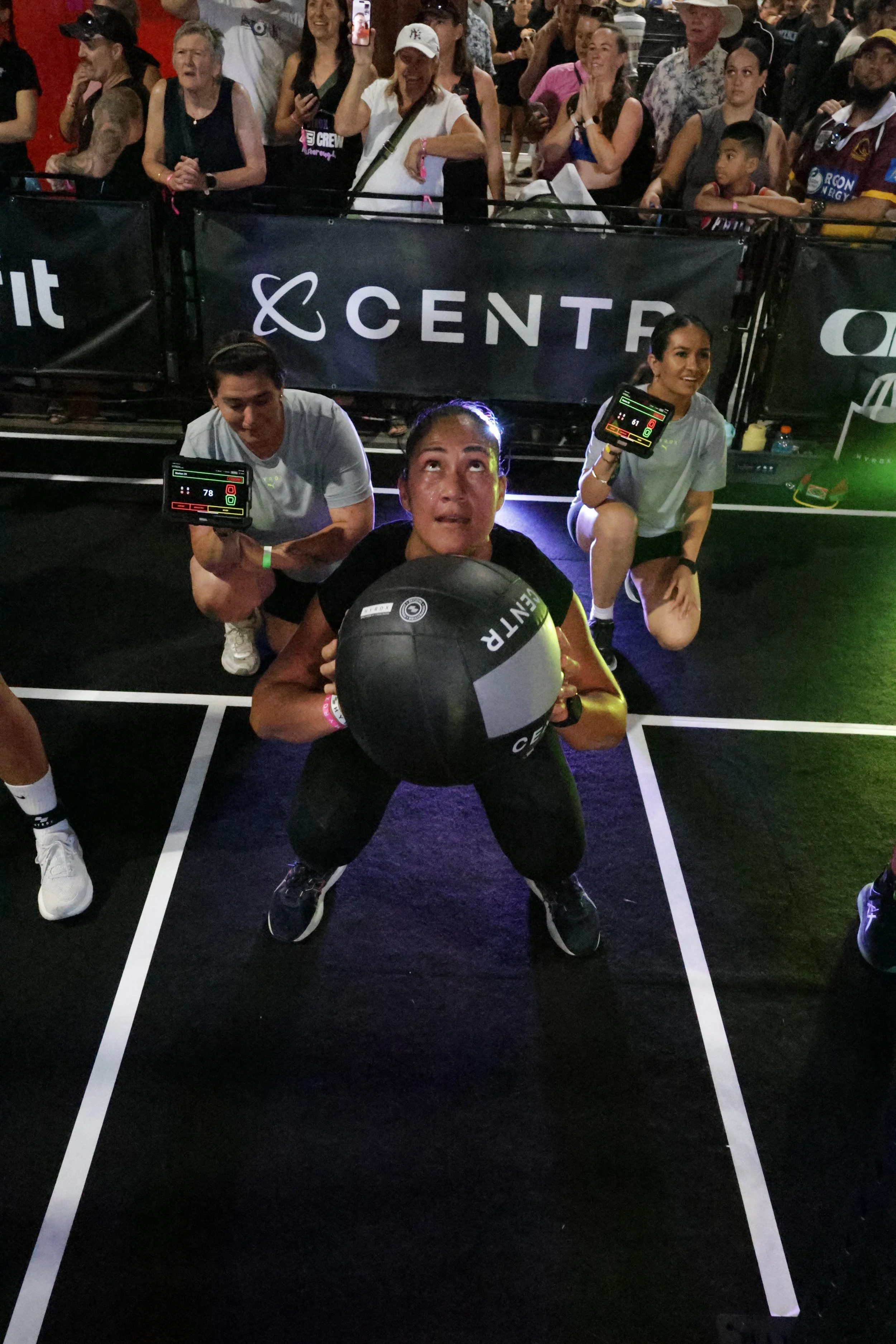 A woman holding a medicine ball in a gym setting with two kneeling women holding electronic tablets, surrounded by a crowd of people watching a fitness event.