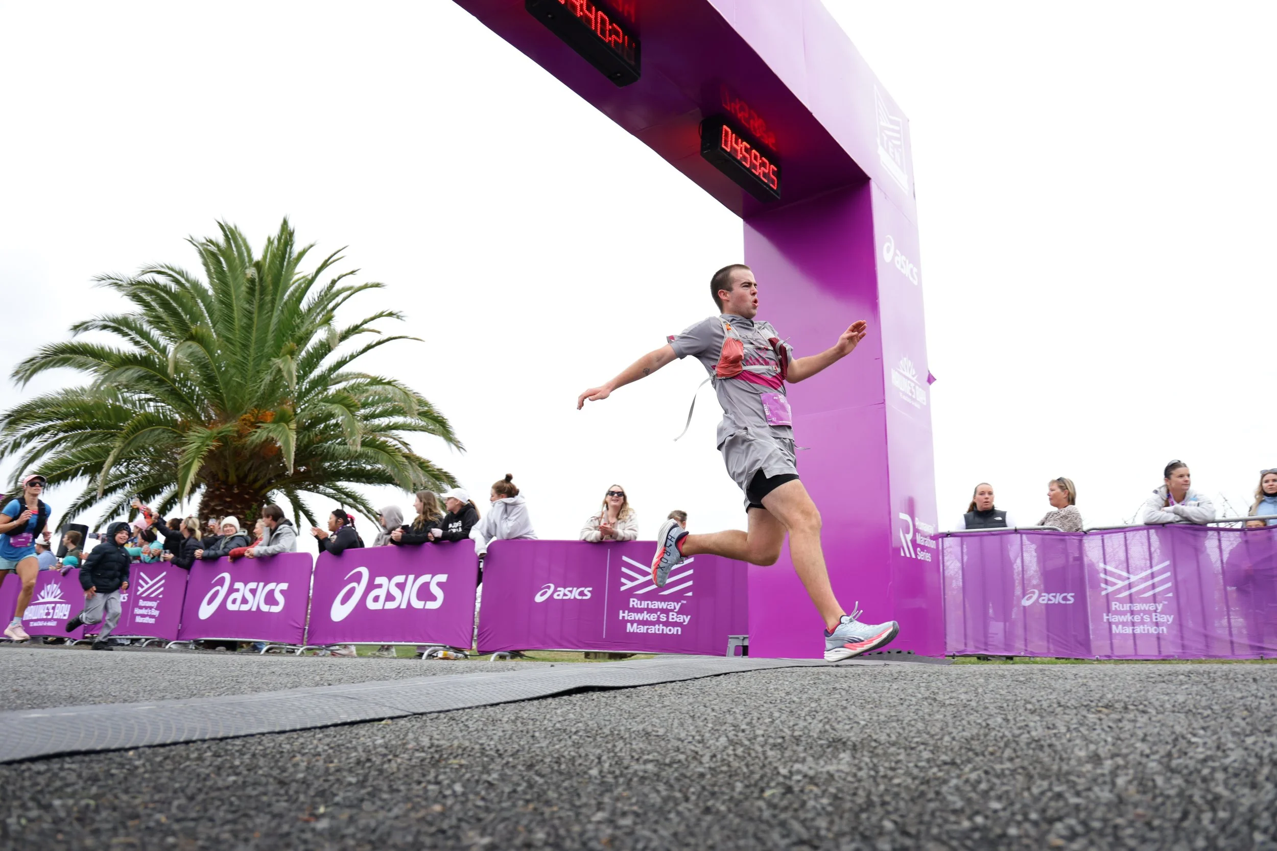 Runner crossing the finish line at the Hawke's Bay Marathon, with a purple finish line arch and spectators behind purple barriers.