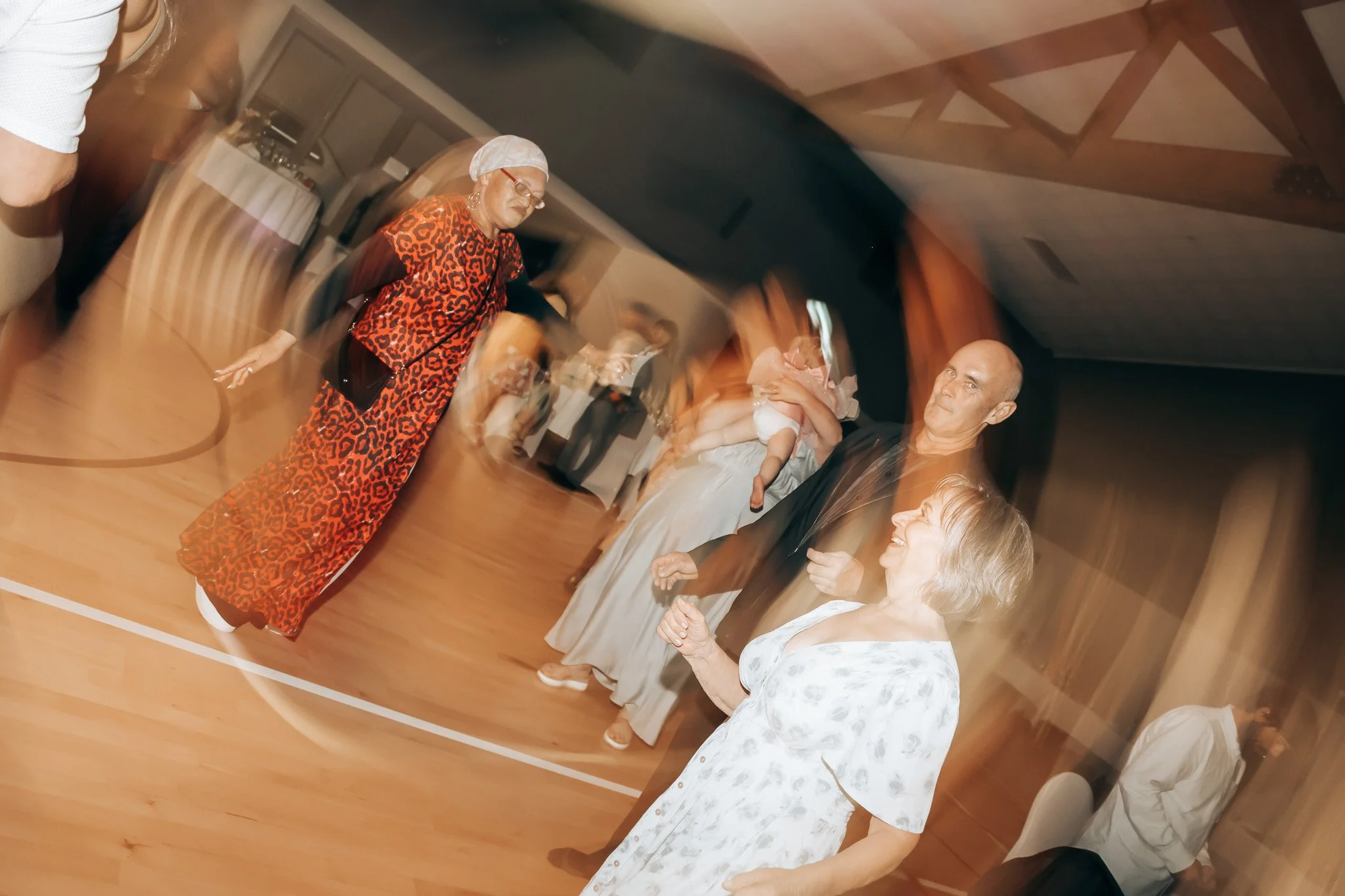A group of people standing indoors in a line, smiling and engaging in conversation, with a woman in a leopard print dress and a head covering in the foreground.