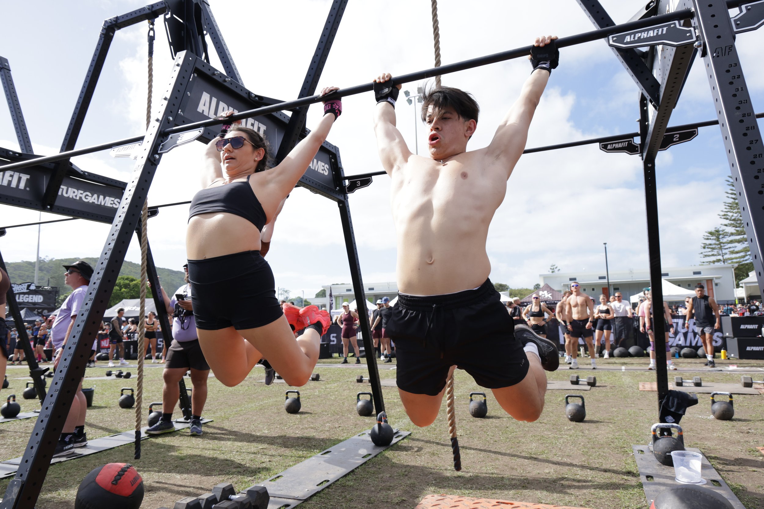 Two people, a woman and a man, are performing a pull-up exercise on a black pull-up bar at an outdoor fitness event. The woman is wearing sunglasses, a black sports bra, and shorts, while the man is shirtless and wearing black shorts. There are other