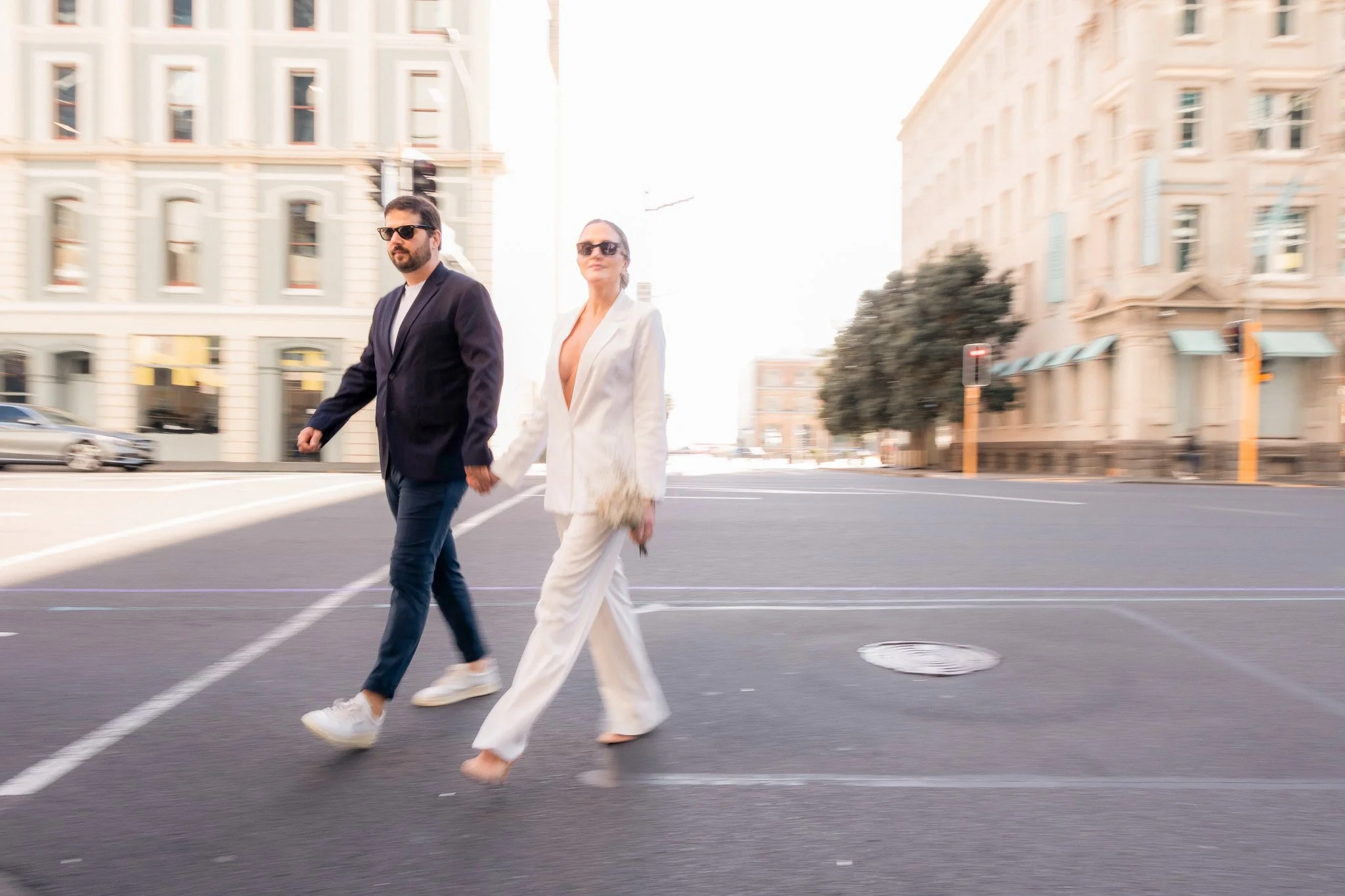A man and woman crossing the street holding hands in an urban area with beige and light green buildings in the background. The man is wearing sunglasses, a dark blazer, and jeans. The woman is dressed in a white suit with a deep V-neck. Both are wear
