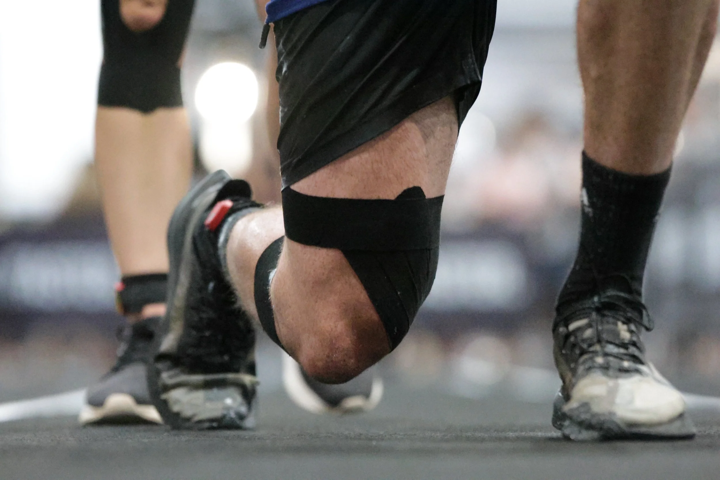 Close-up of a person in black athletic shorts and black knee-high socks, crouching in a workout or sports setting, with other individuals visible in the background.