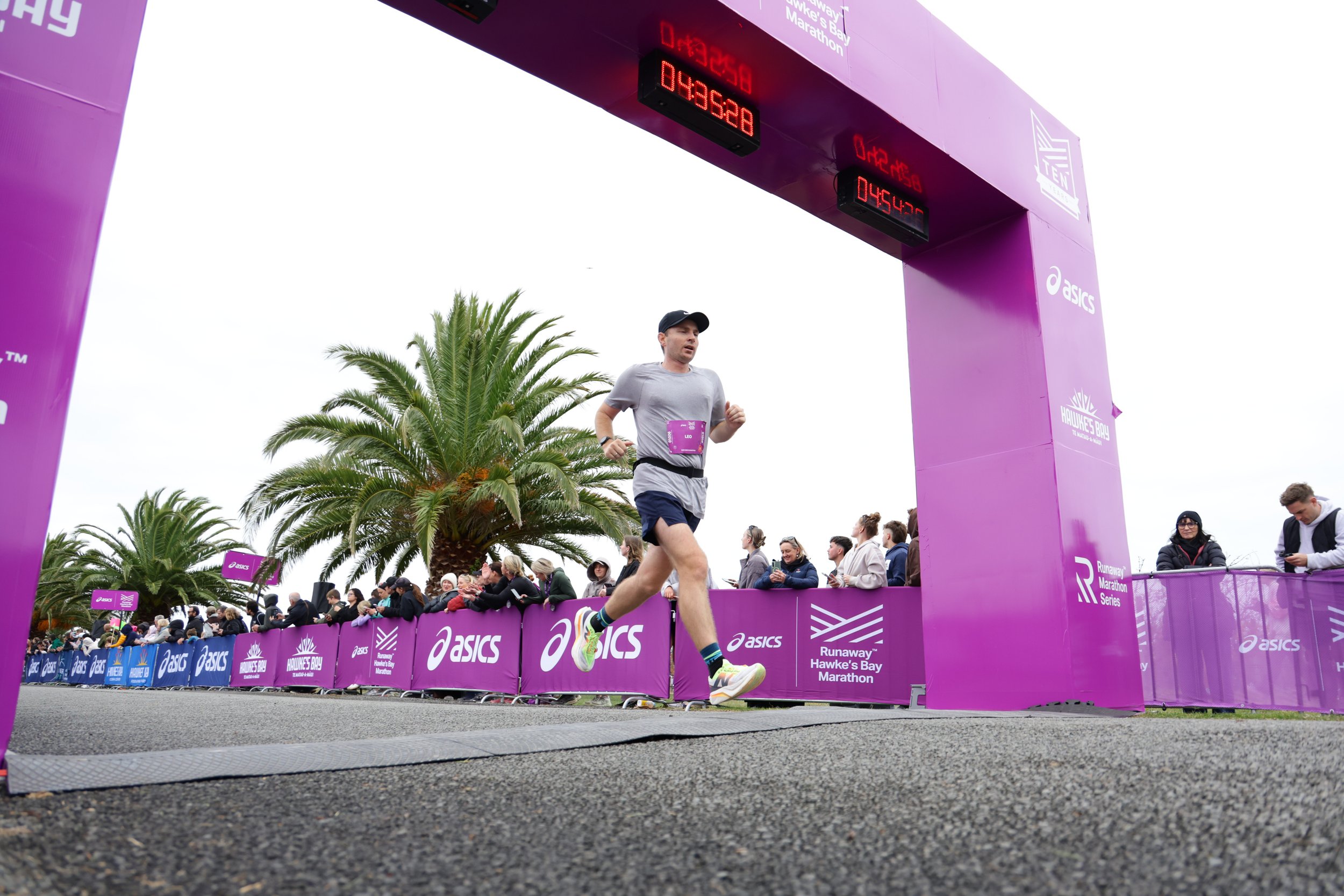A man running in a marathon with people watching behind a purple barrier, under a purple archway with digital clocks and sponsorship logos. Palm trees are visible in the background.