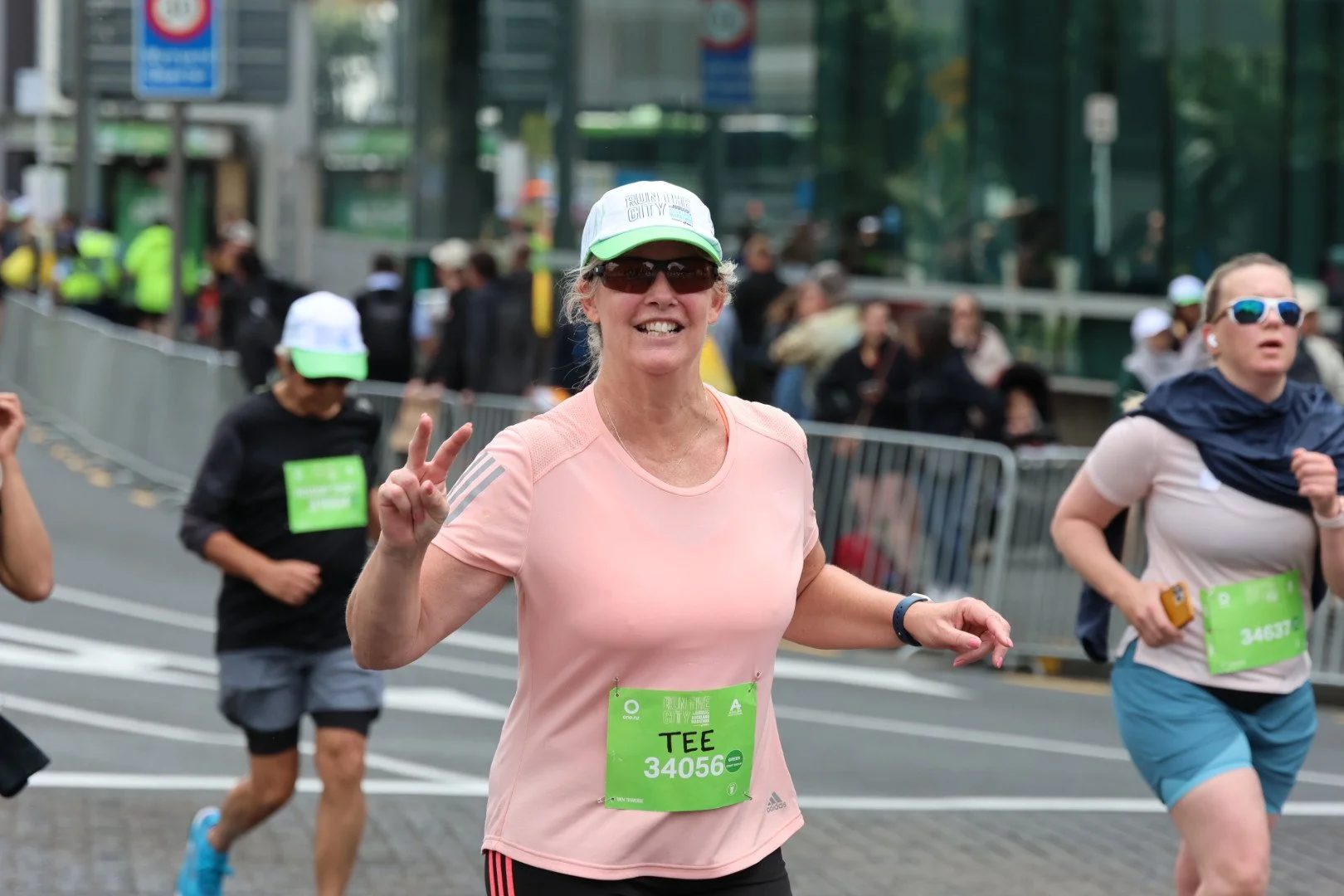 Happy woman with sunglasses and a white cap making a peace sign while running in a marathon, with other runners and spectators in the background.