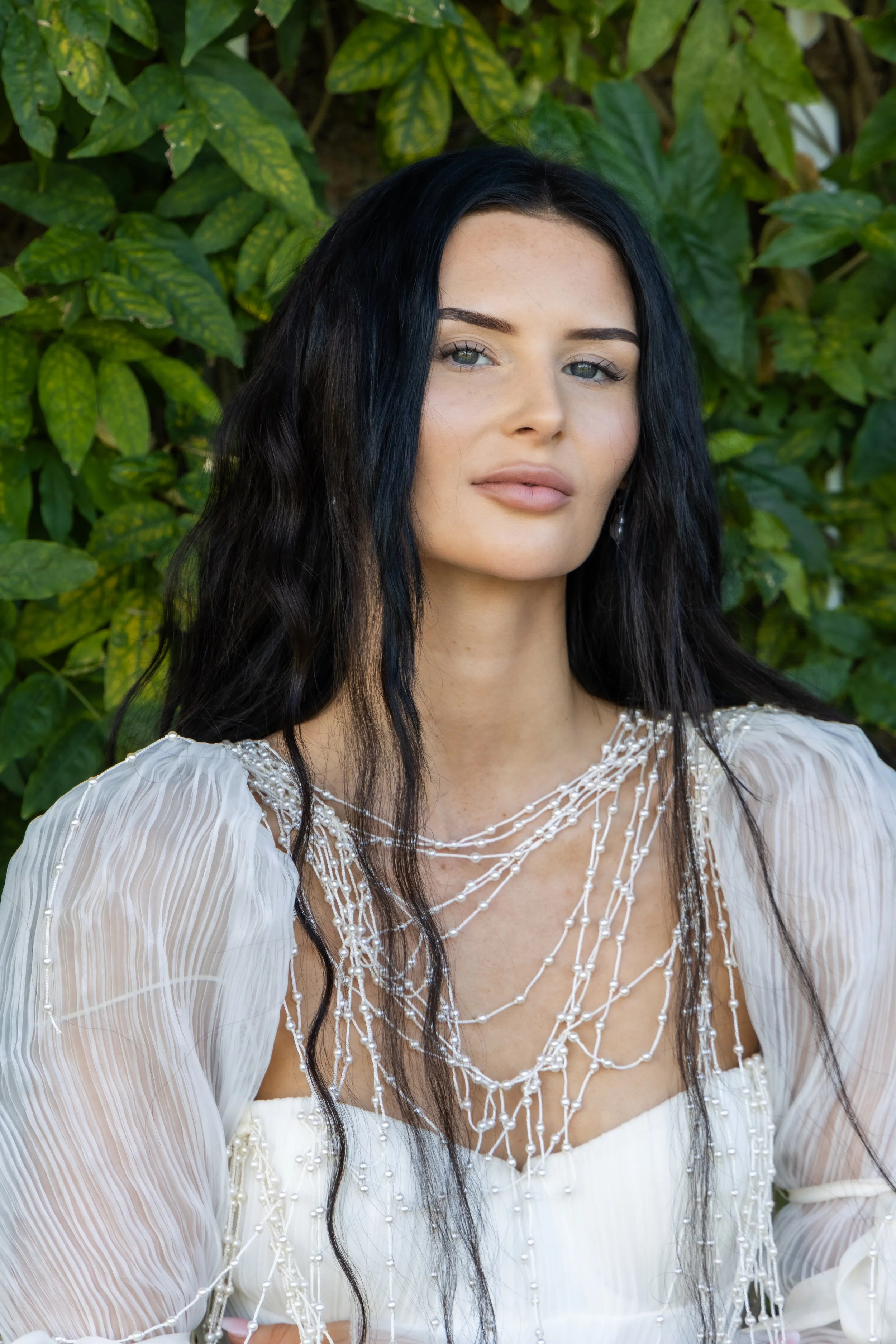 A woman with long black wavy hair, wearing a white, sheer, beaded dress with pearl details and pearl earrings, standing in front of a green leafy background.