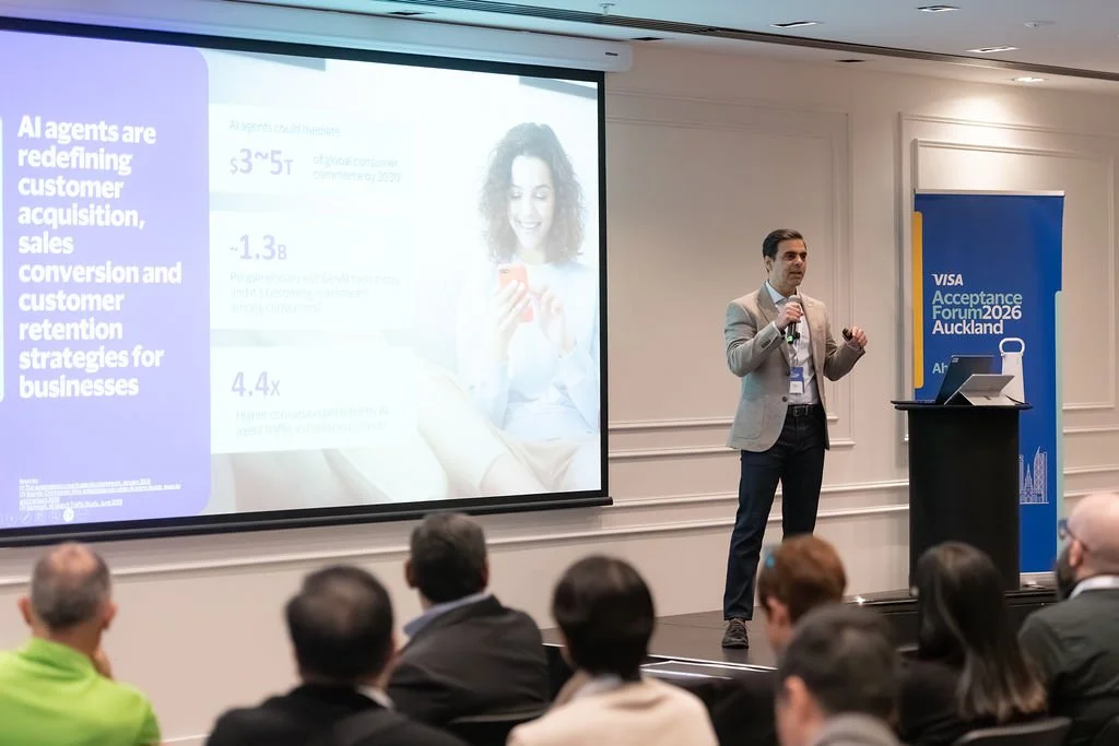 A man in a gray blazer giving a presentation at the Visa Acceptance Forum 2026 in Auckland, standing on stage with a microphone, in front of a large screen with a slide, while audience members watch.