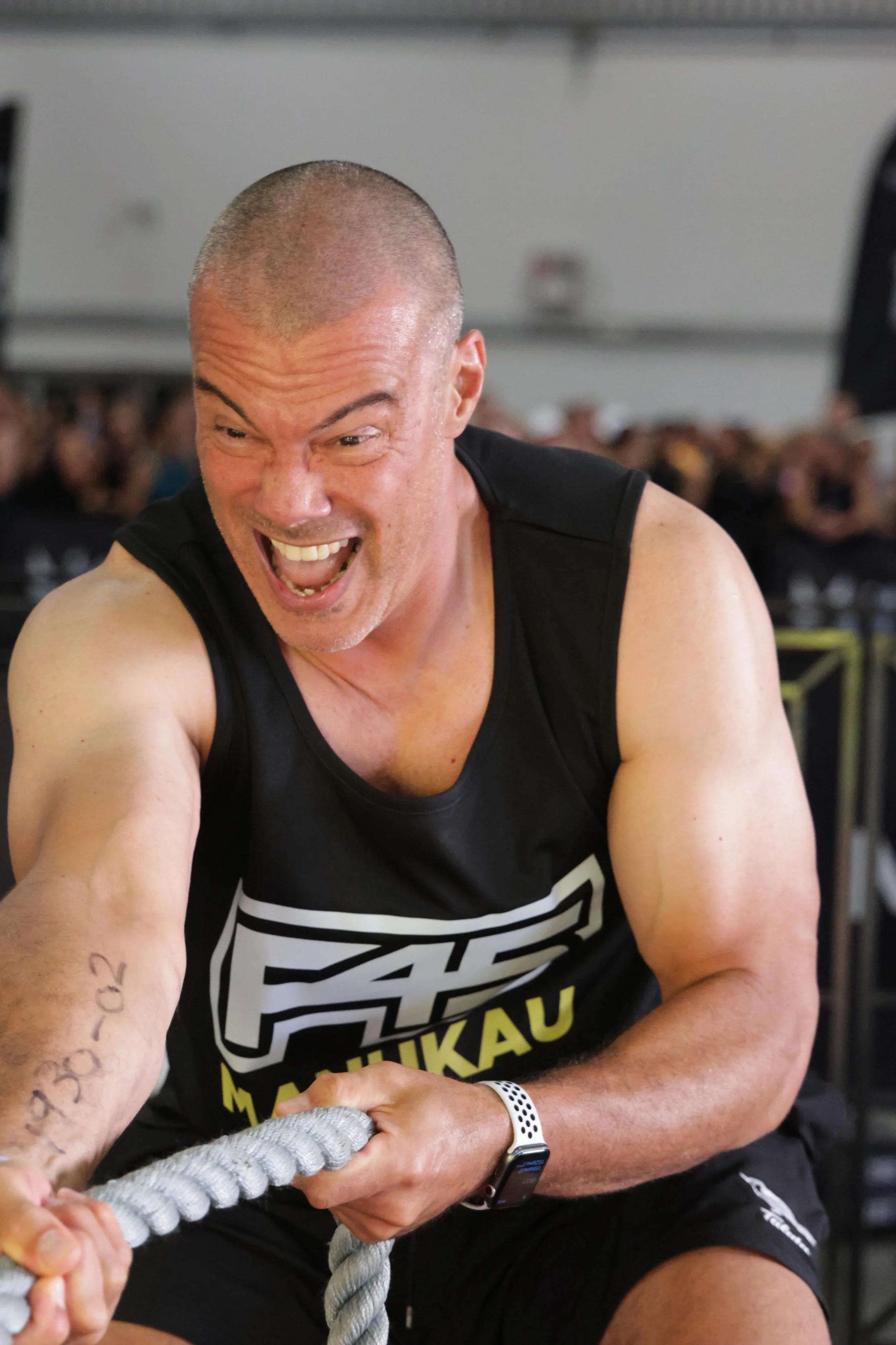 A man with a shaved head and tattoos on his arm, wearing a black tank top with branding and a smartwatch, is participating in a fitness activity involving pulling on a thick rope in a gym filled with people.