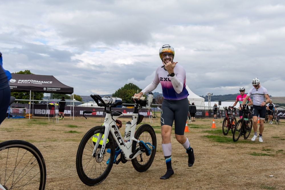A woman wearing a white and purple triathlon suit, helmet, and sunglasses is smiling and making a thumbs-up gesture while standing next to her white Felt bicycle at an outdoor triathlon event. Other participants are in the background, some with bikes