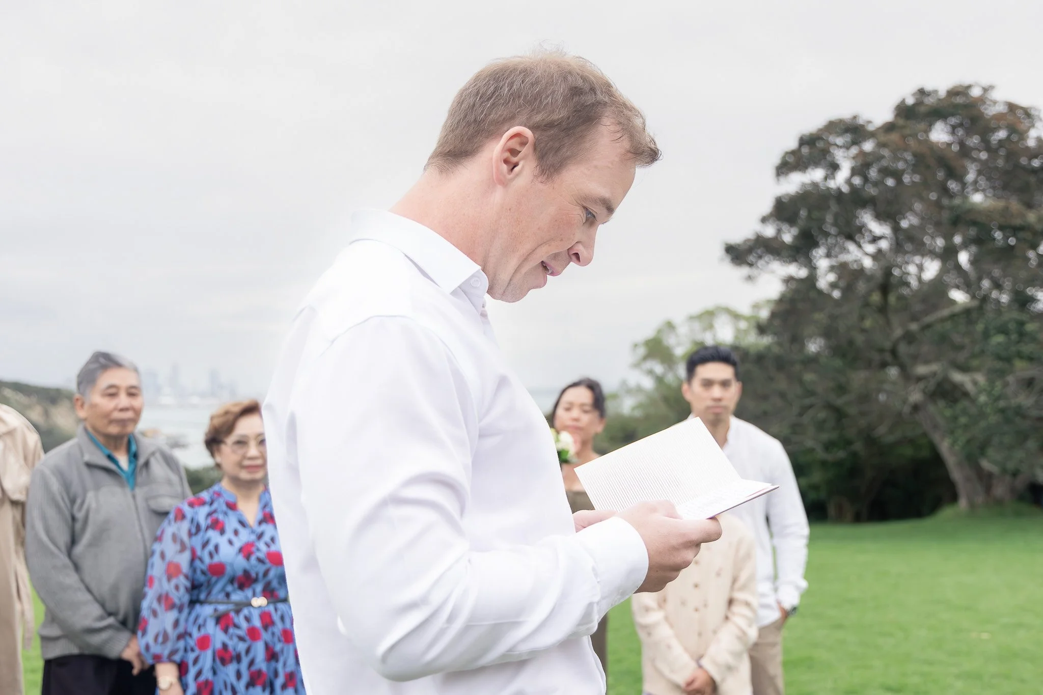 A group of people gathered outdoors, with a man in a white shirt reading from a paper, possibly participating in a ceremony or event, with trees and a cloudy sky in the background.