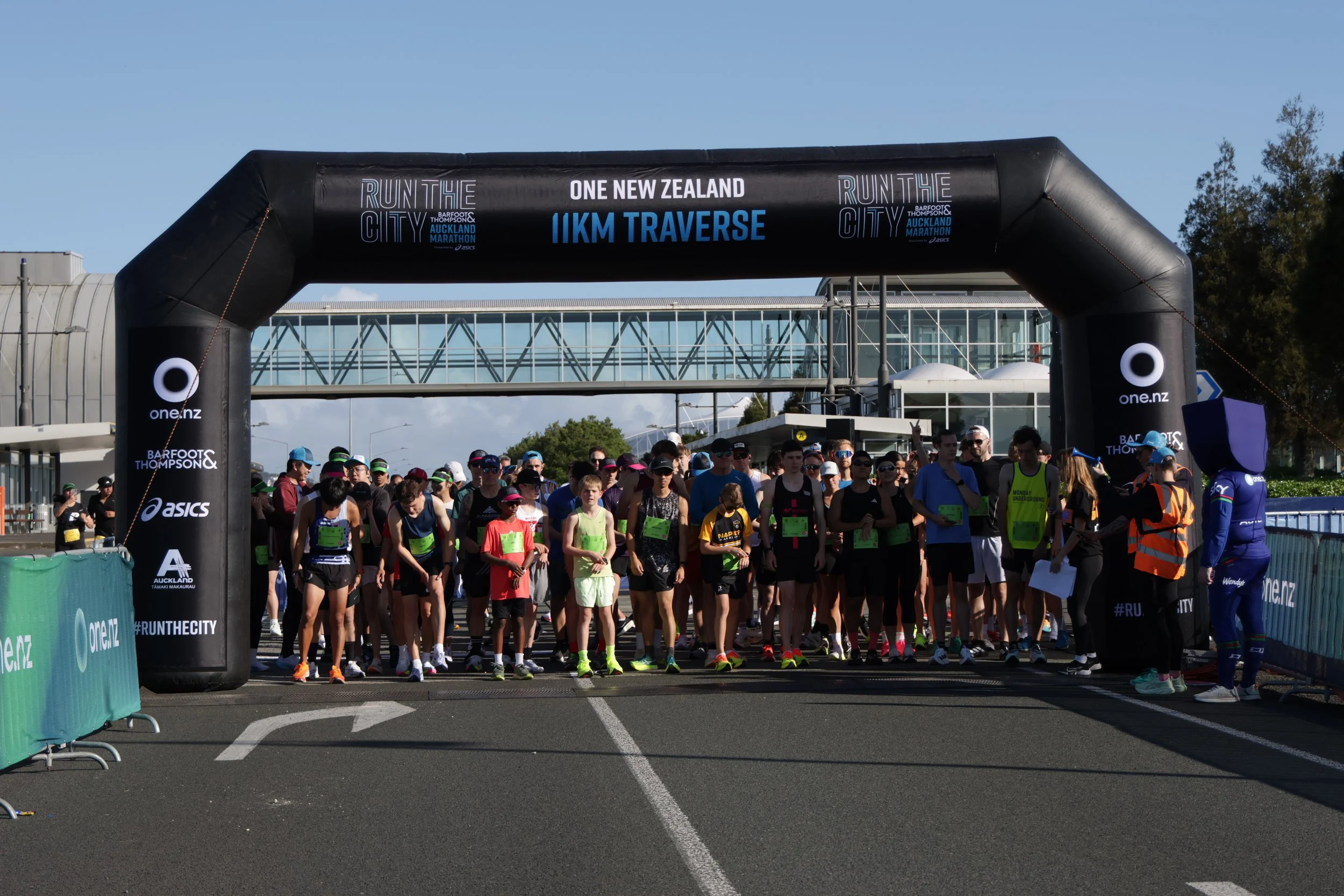 Start line of a marathon race with many runners gathered beneath a large black inflatable archway that reads 'Run the City, One New Zealand, 11KM Traverse' with a blue sky and some trees in the background.