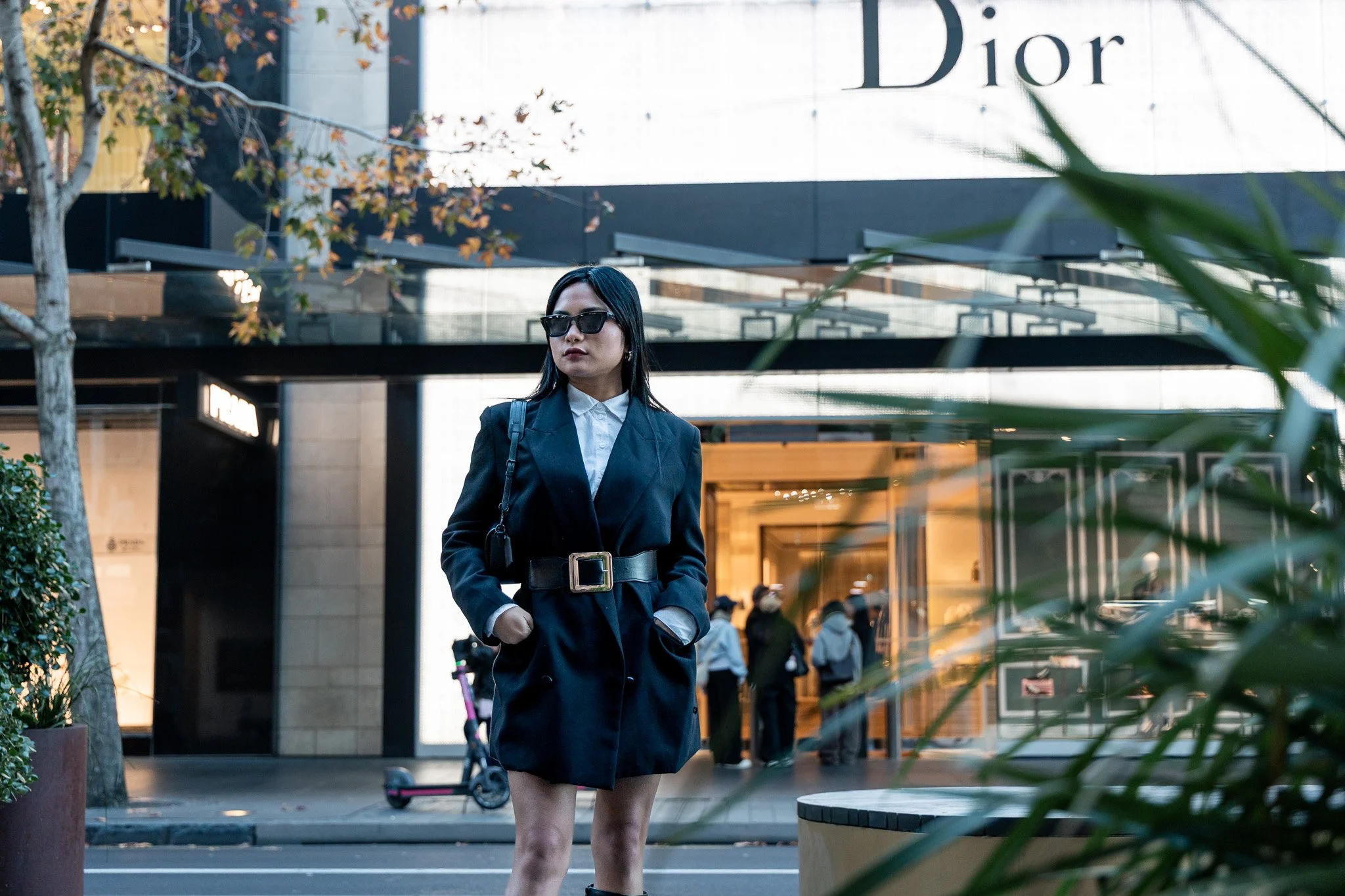 A stylish woman in a black blazer and sunglasses walking outside near a Dior store, with a scooter and a crowd in the background.
