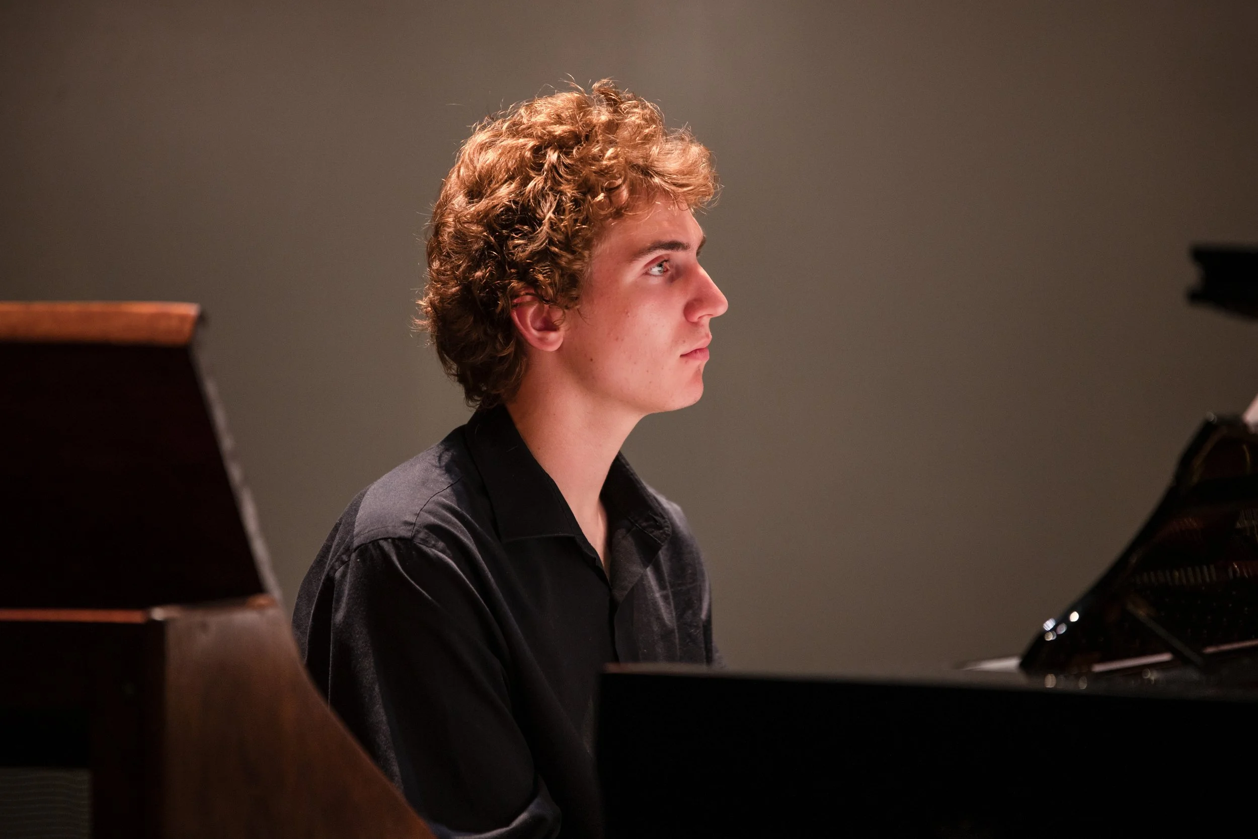 A young man with curly blonde hair and a serious expression playing the piano, dressed in a black shirt, in a dimly lit room.