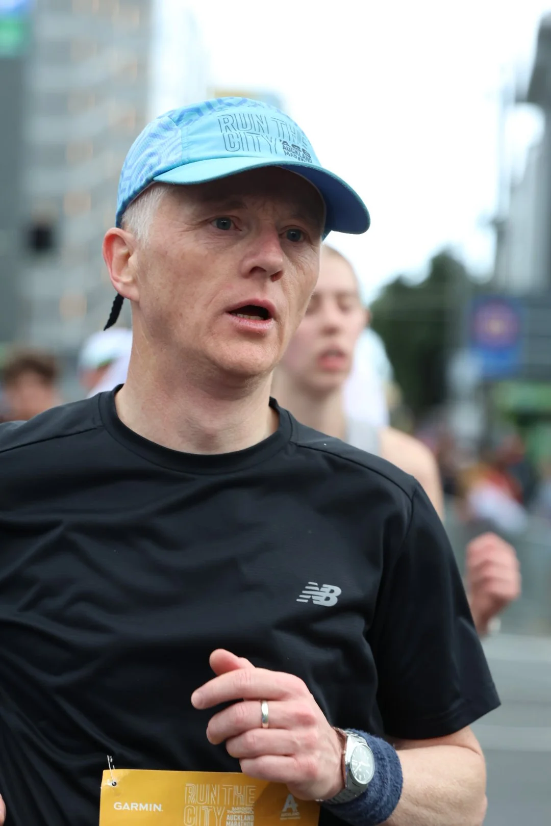 Man running in a race wearing a black athletic shirt and a blue cap, with a focused expression, and a yellow race bib pinned to his shirt.