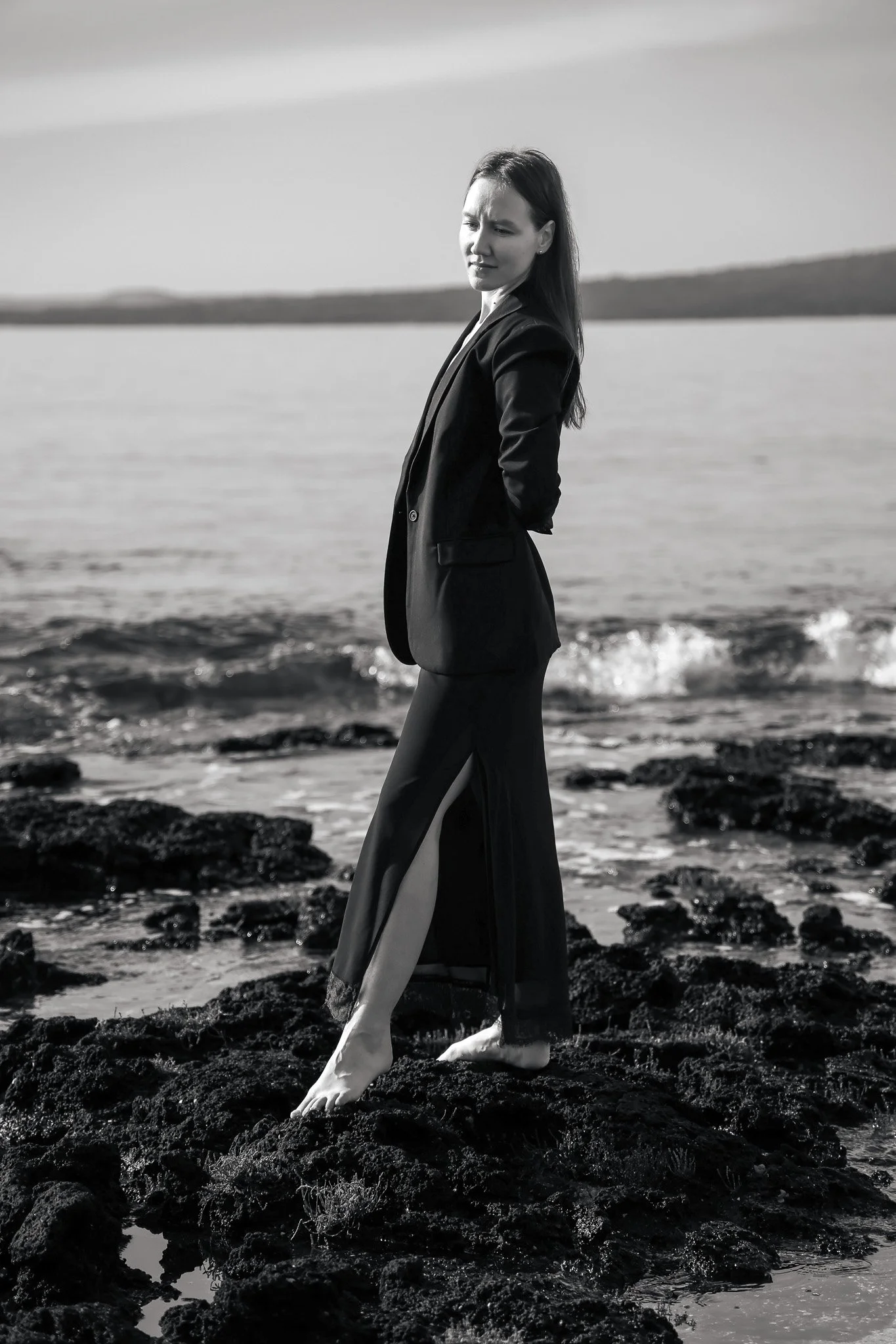 A woman in a black suit coat and colorful patterned dress with high slit, standing barefoot on rocks at the shoreline with water and land visible in the background, in black and white.