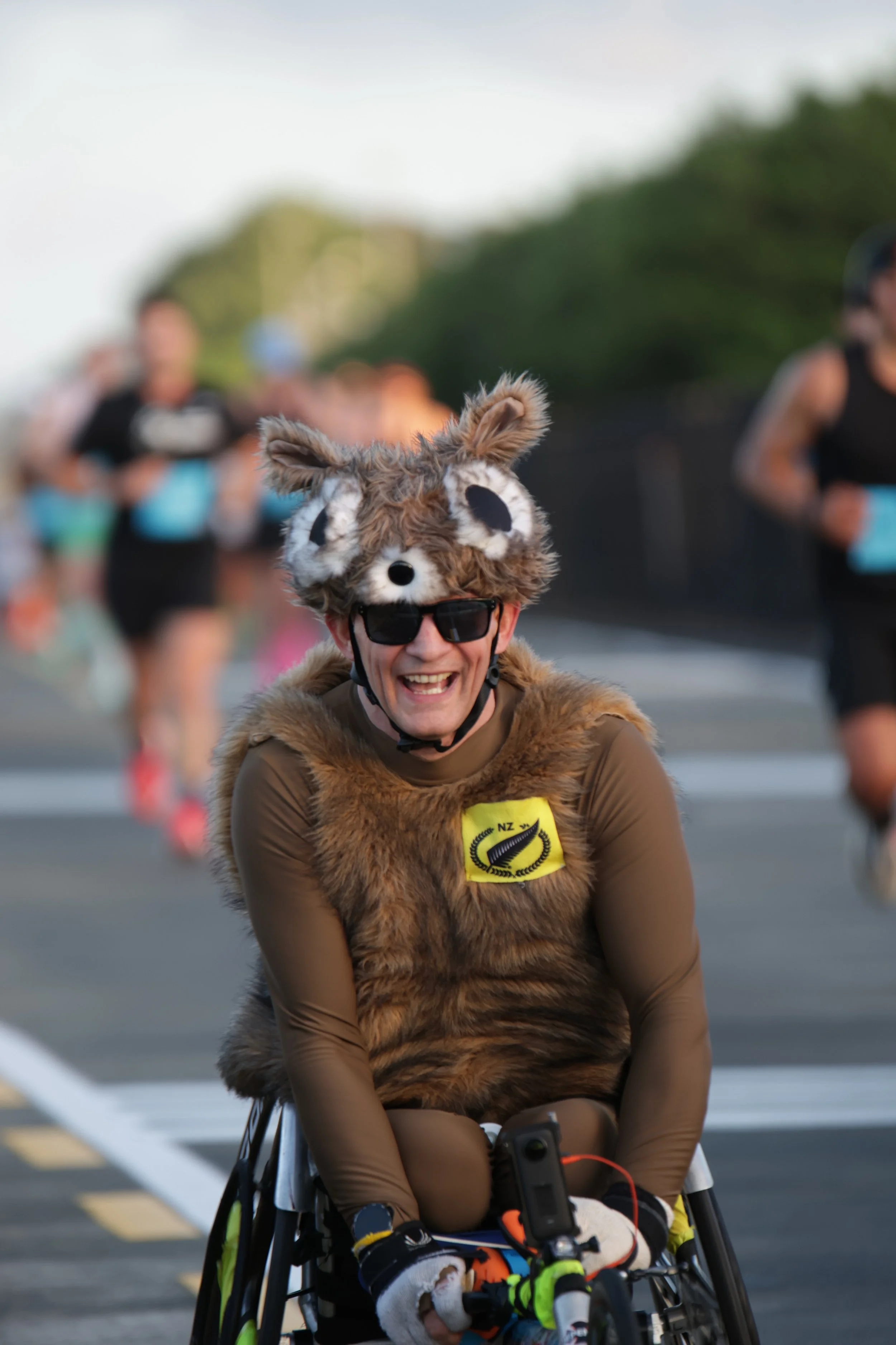 Person smiling in a wheelchair at a race, wearing a costume with a wolf hat and furry vest.