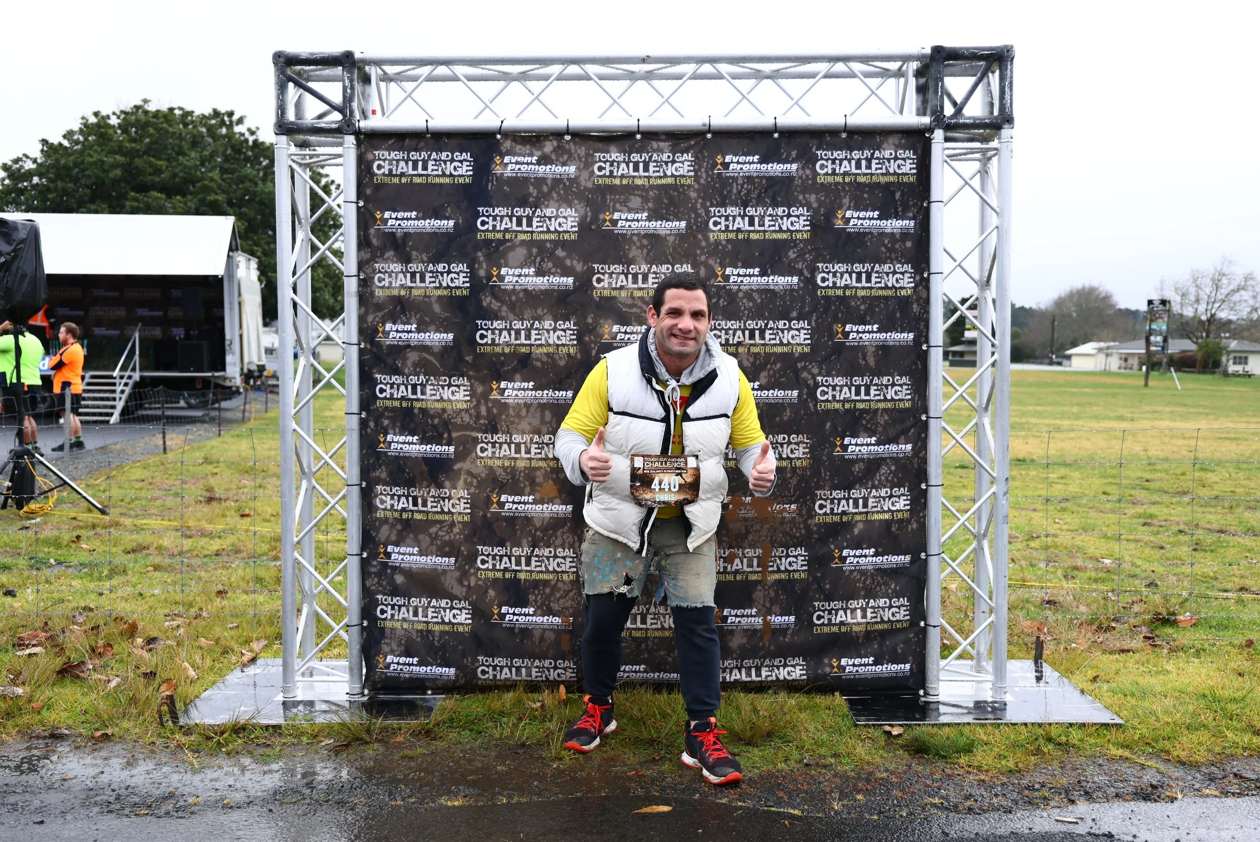 A man in a gray vest and yellow shirt giving a thumbs-up pose with mud on his clothes, standing in front of a black backdrop with event logos for the Tough Guy and Gal Challenge, an off-road running event.