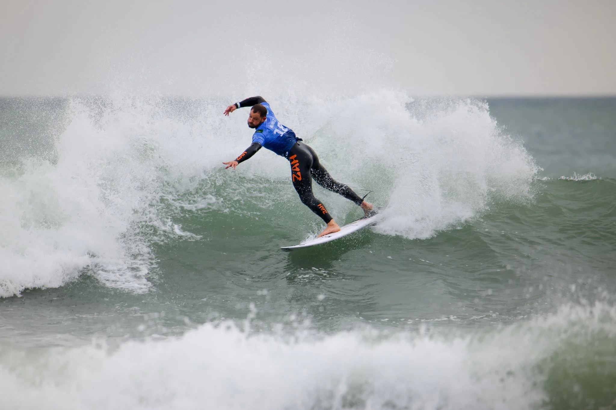 A male surfer wearing a wetsuit and a blue jersey riding a wave on a surfboard in the ocean.