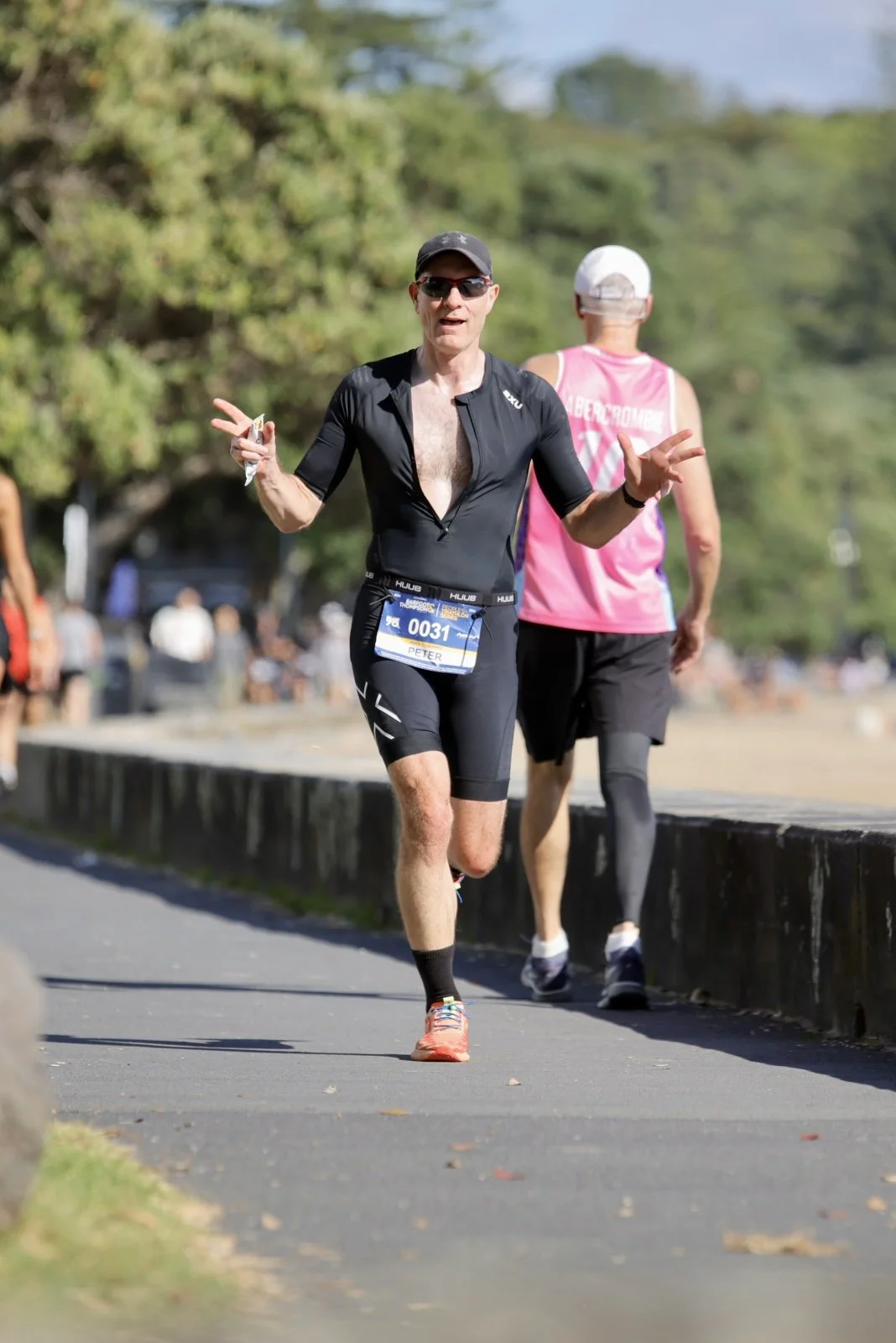 A man running in a marathon or race, wearing sunglasses, a black athletic outfit, and a race bib with the name Peter, with trees and other runners in the background on a sunny day.