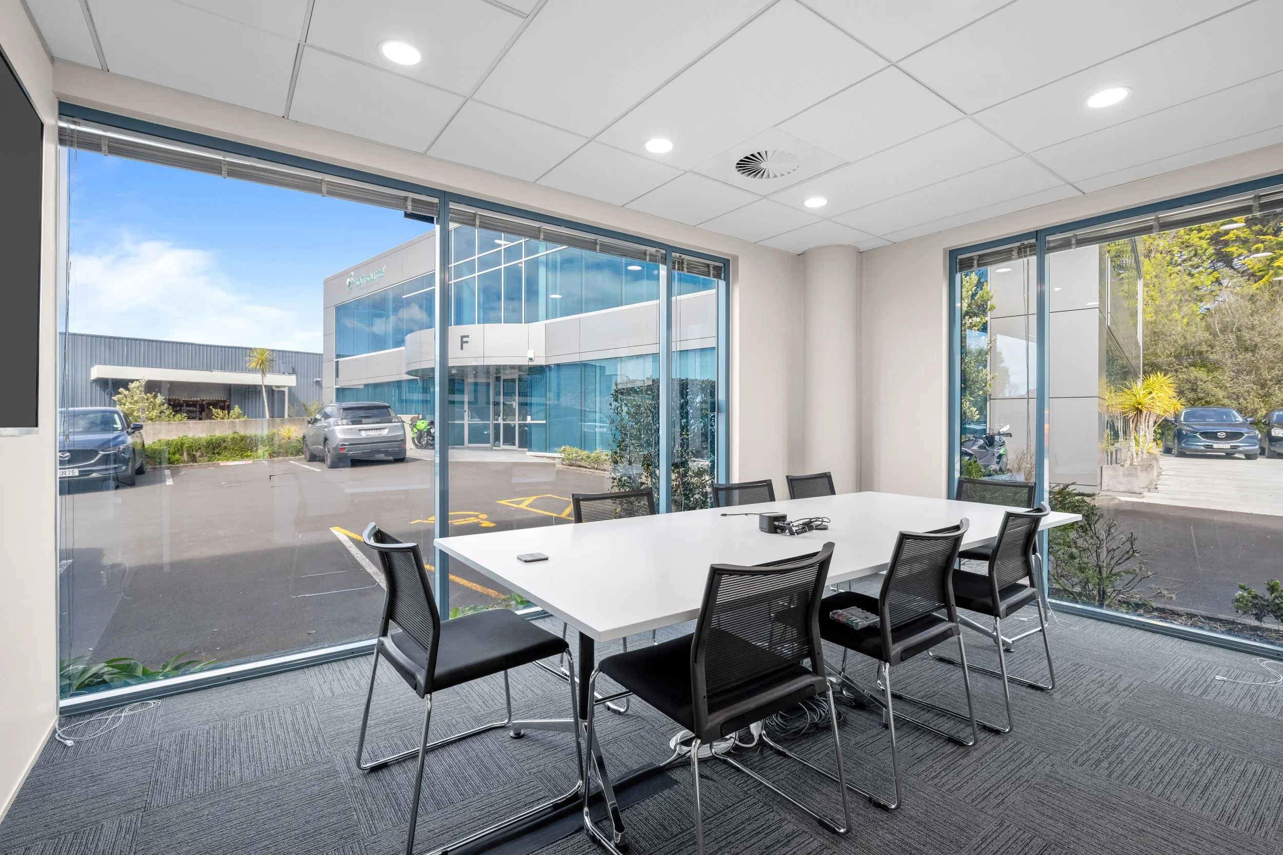 A conference room with large glass windows showing a parking lot and modern office buildings outside. Inside, there is a white table surrounded by black mesh office chairs, and some electronic devices are placed on the table. The ceiling has recessed