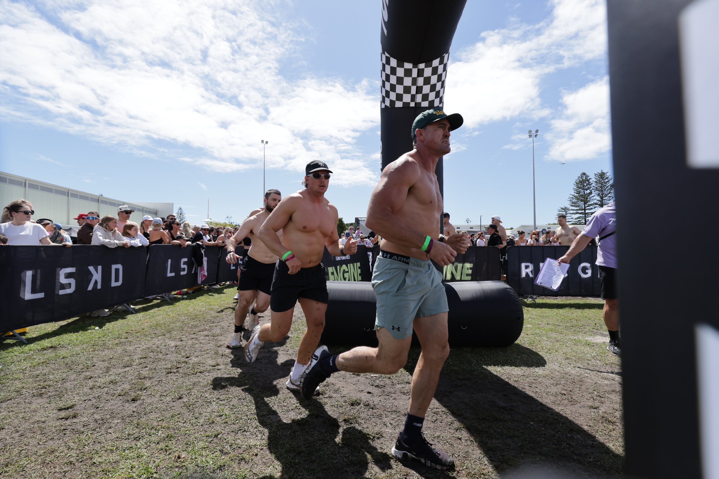 Men running in a race, emerging from a black inflatable arch with checkered pattern, with a crowd of spectators behind a black barrier, under a partly cloudy sky.
