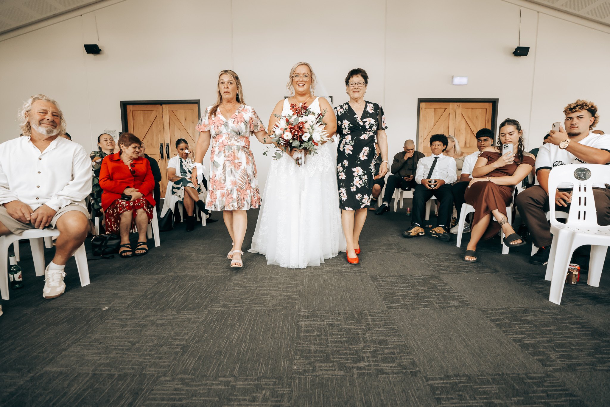 A bride in a white wedding dress holding a bouquet, walking down the aisle with two women, surrounded by seated guests in a large, indoor venue.