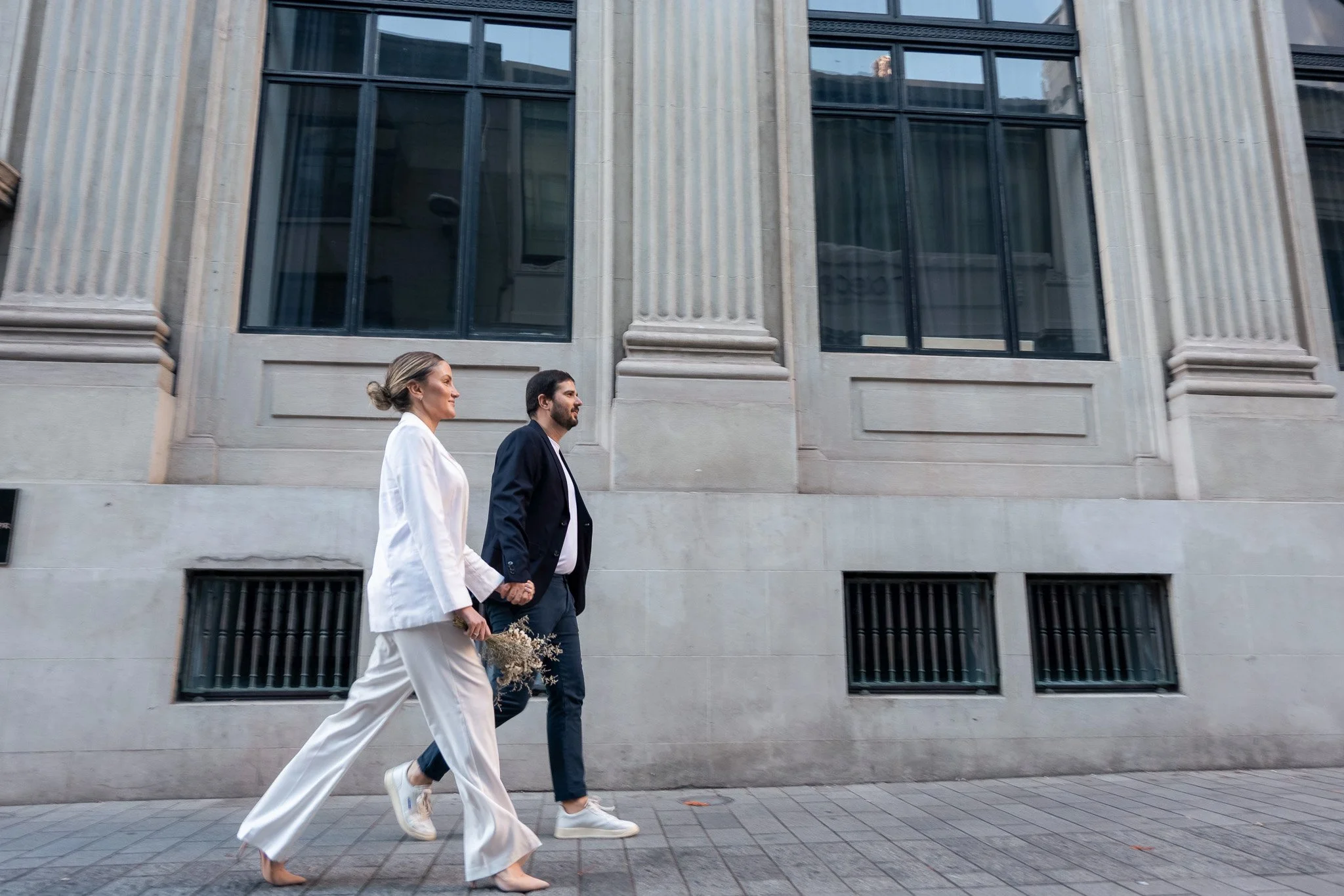 A woman in a white suit and a man in a dark suit walking together on the sidewalk in front of a building with large windows and columns.