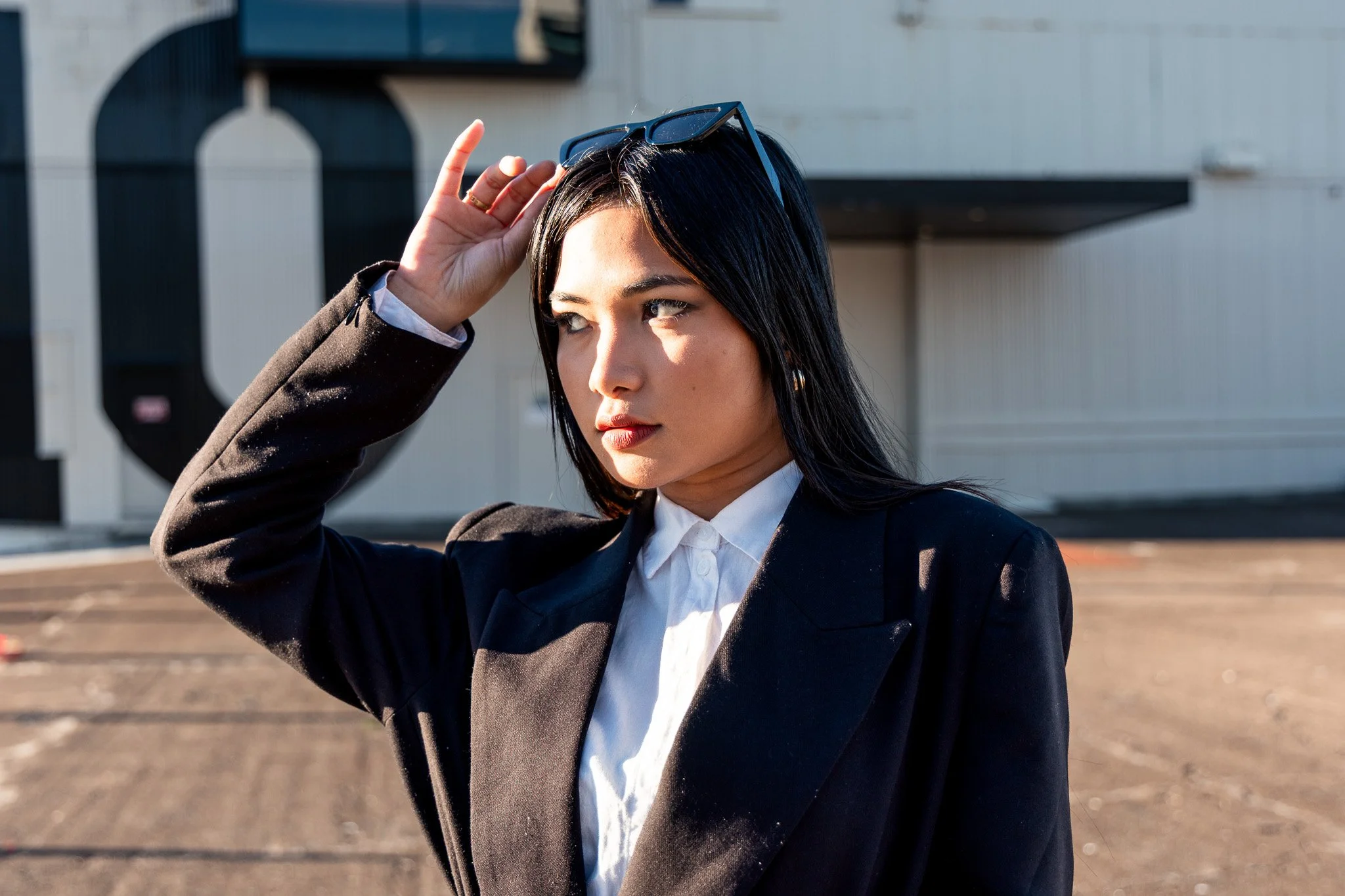 A woman in formal attire adjusting sunglasses on her head outdoors with a building and parking lot in the background.