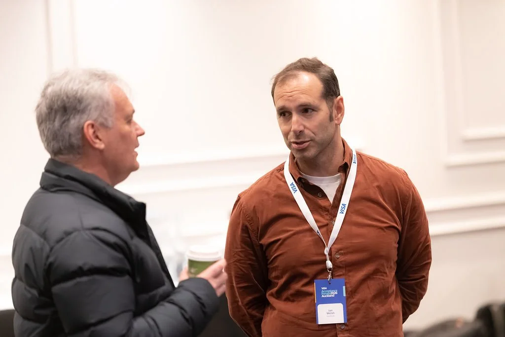 Two men engaging in conversation at a conference, with one holding a coffee cup and wearing a black jacket, and the other wearing a brown shirt with a conference badge around his neck.