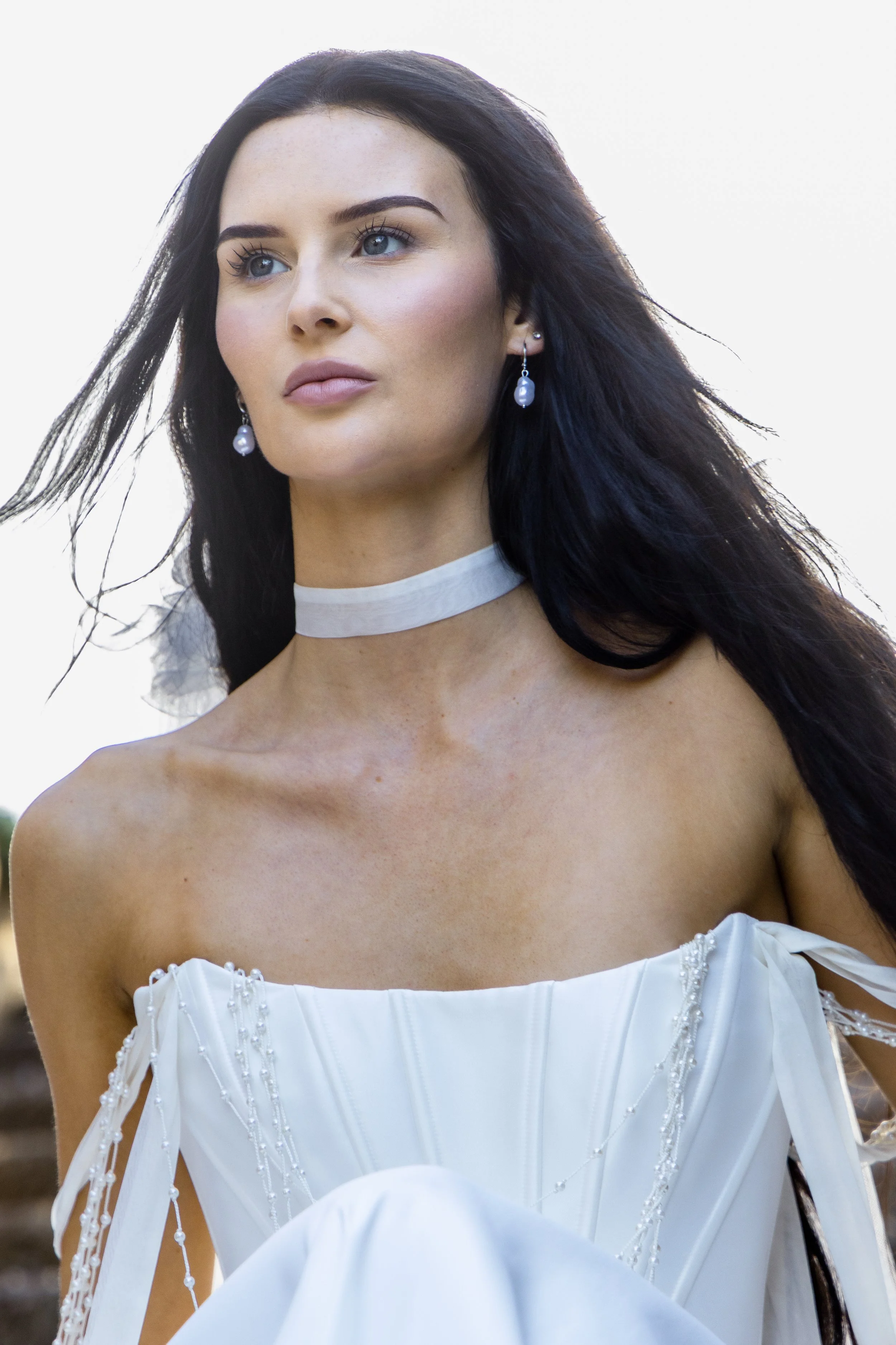 A woman with dark hair, blue eyes, and pearl earrings posing outdoors in a white dress with decorative beads and ribbons.