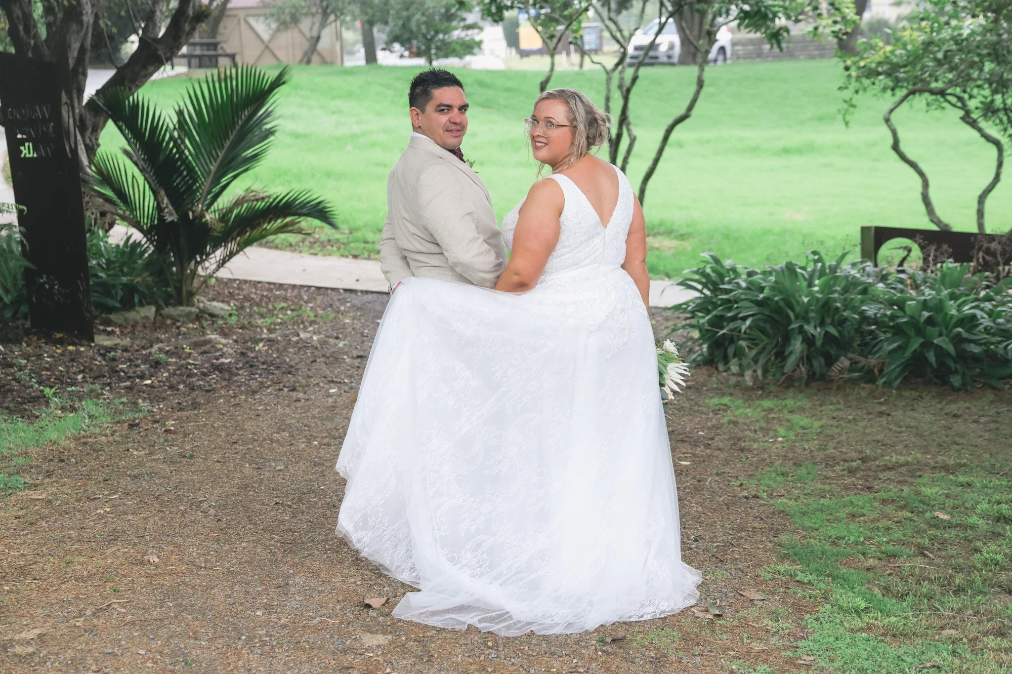 A newlywed couple in wedding attire standing outdoors in a lush green park. The bride is wearing a white lace wedding dress with an open back and glasses, and the groom is in a light-colored suit. They are holding hands and looking back at the camera