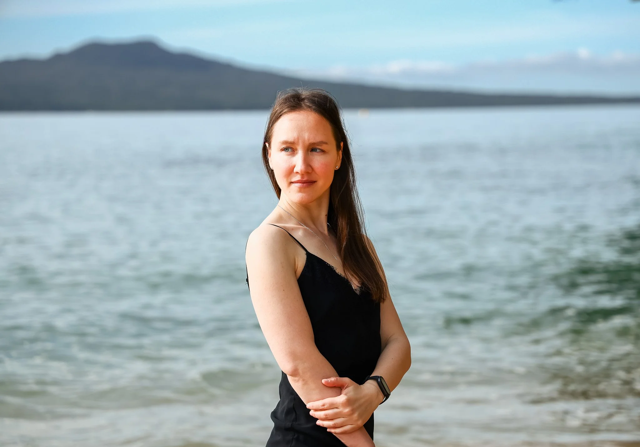 A woman in a black top standing at the beach, with calm water and a distant mountain in the background.