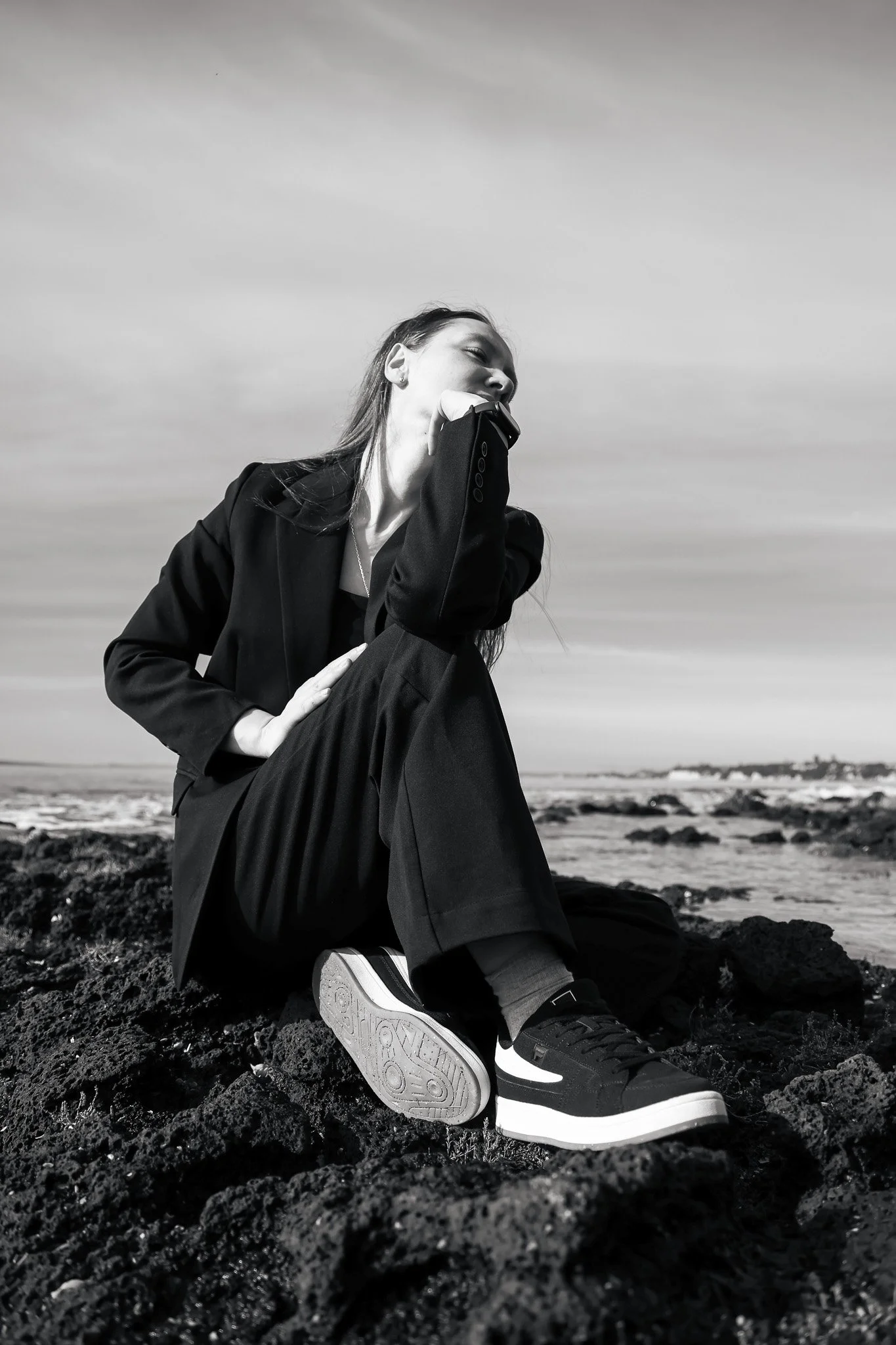 A black and white photograph of a woman sitting on rocks near the water, with her head tilted back and eyes closed. She is dressed in a dark blazer, pants, and sneakers, with one hand resting on her knee and the other raised to her face. The landscap