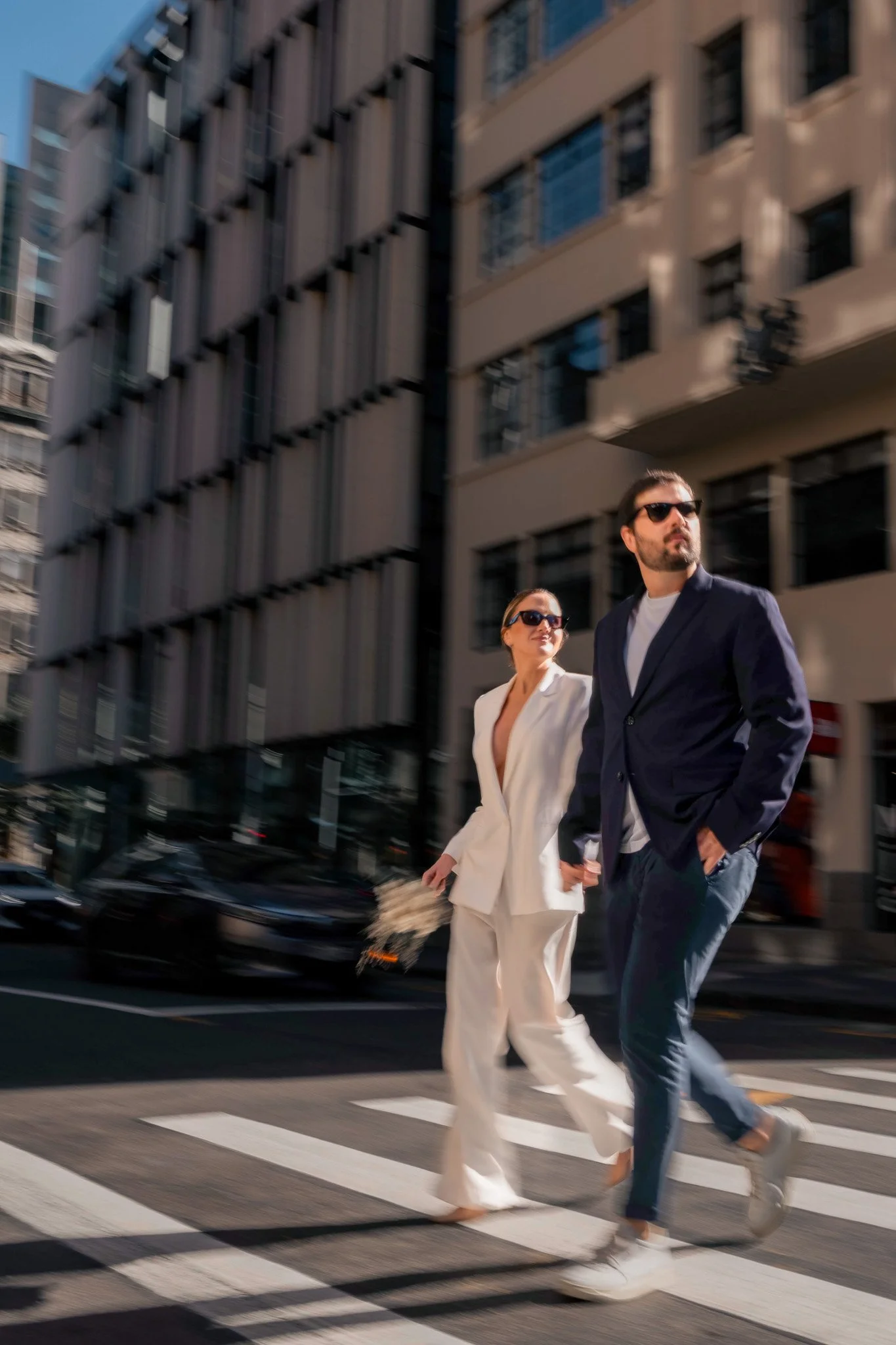 A man and woman are walking across a city crosswalk in business attire, with blurred buildings and cars in the background.