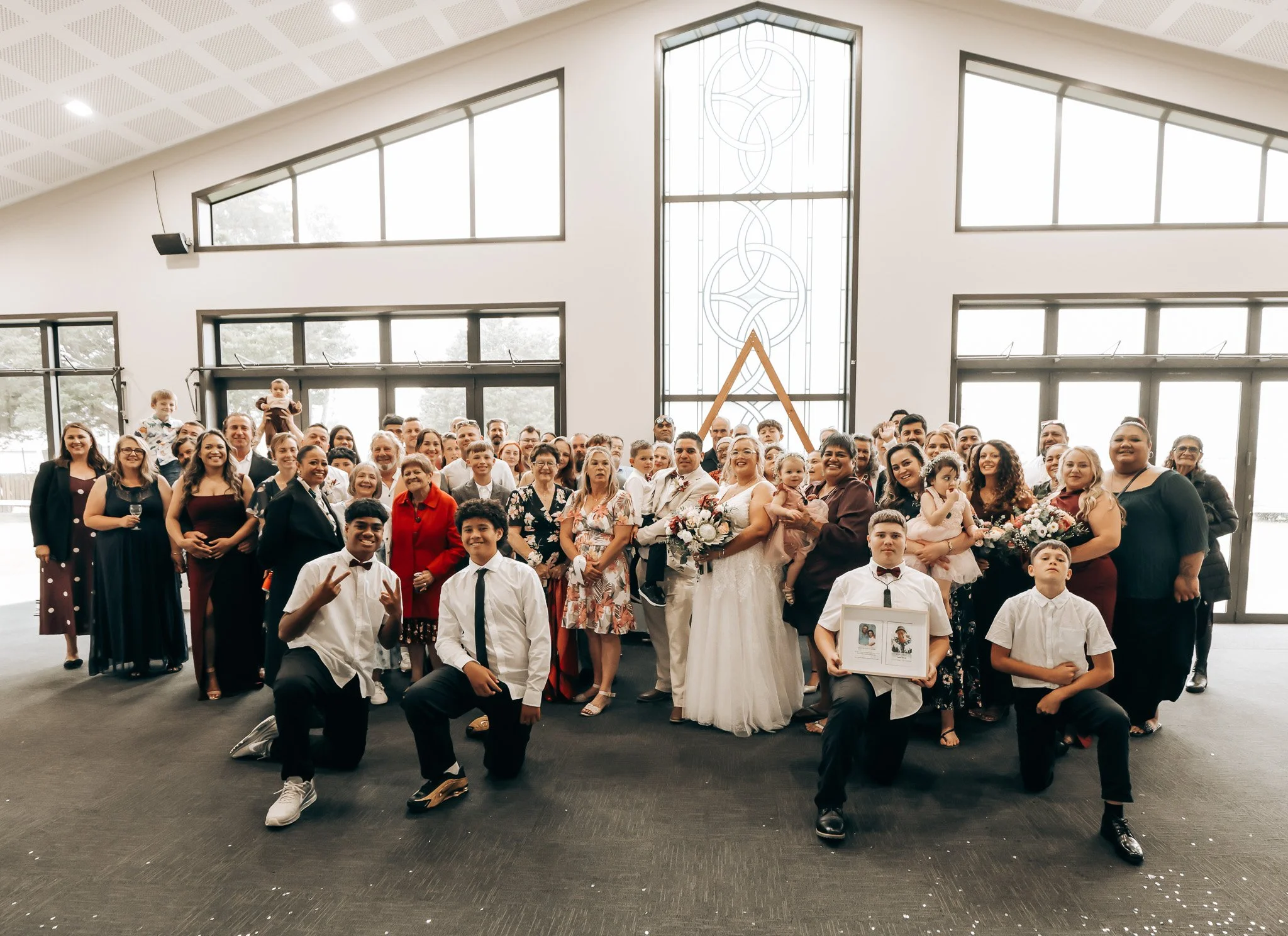 A large group of men, women, and children dressed in formal attire gathered inside a church for a wedding celebration, with some holding flower bouquets and framed photos, and a large stained glass window in the background.